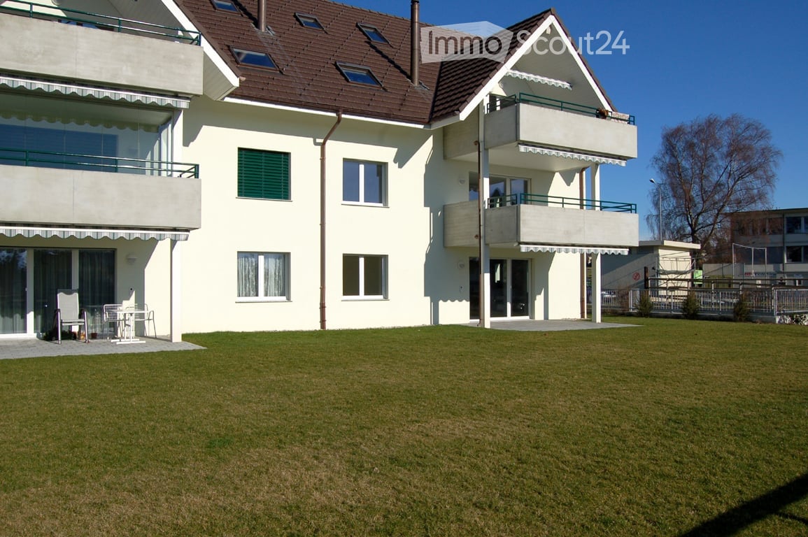 Two-story house with white walls, brown roof, multiple windows, and balconies. A small patio area with a table and chairs is visible on the left side.