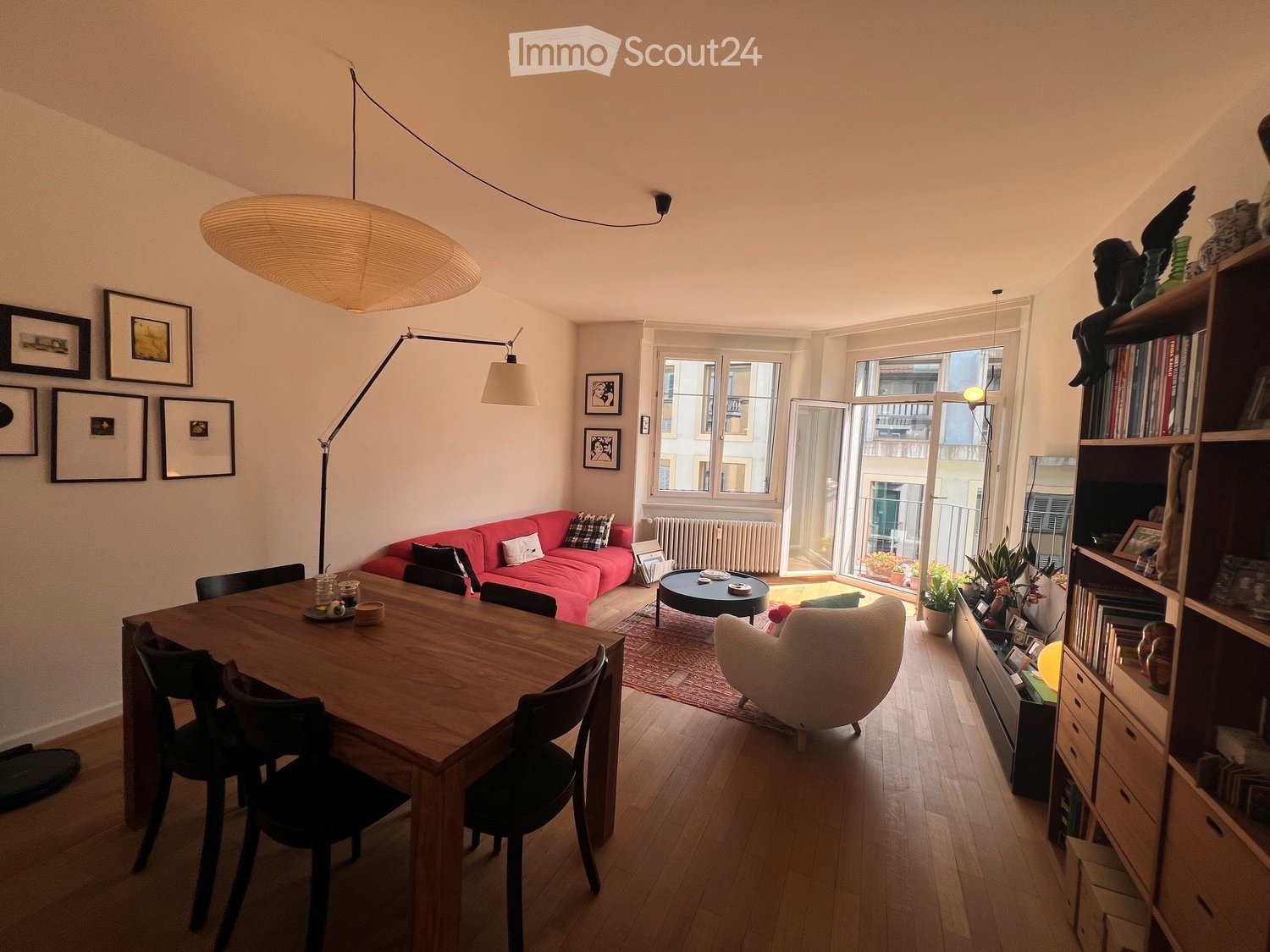 Bright living and dining area with wooden flooring, large windows, a red sofa, a wooden dining table with chairs, a bookshelf, and decorative items.