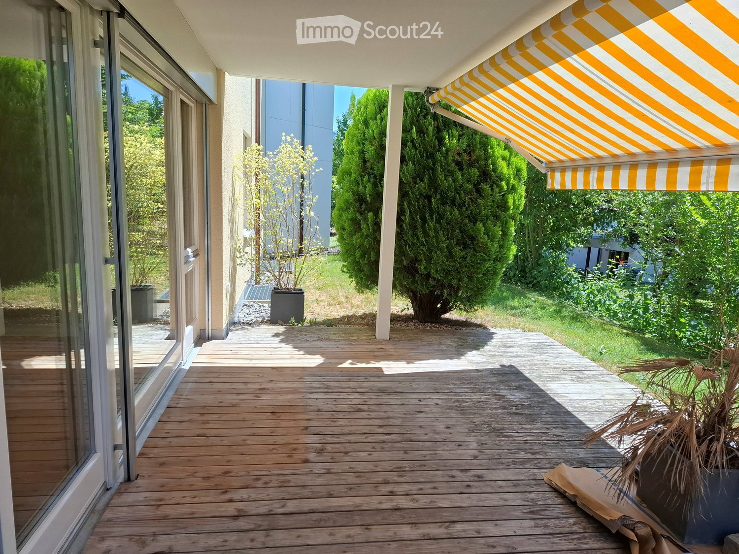 Wooden deck with a retractable awning, large windows, and a view of a green garden with trees and shrubs.