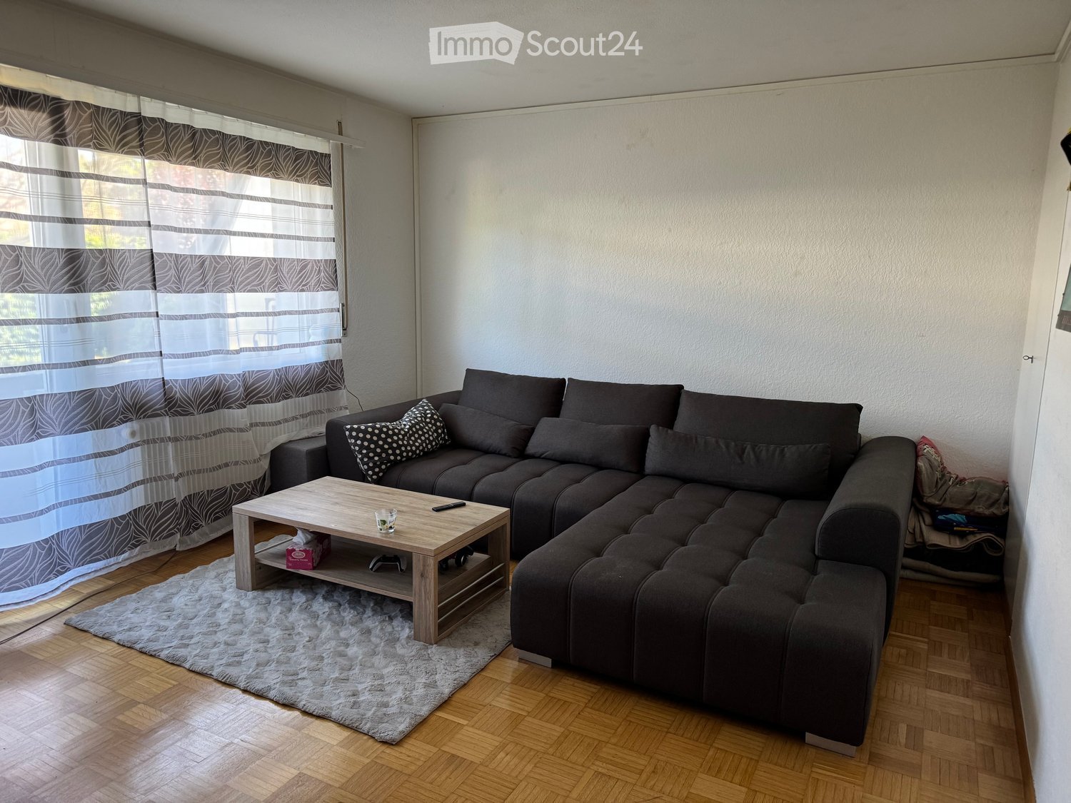 Modern living room with a dark grey sectional sofa, wooden coffee table, and patterned throw pillow. The room features wooden flooring and a large window with grey and white curtains.