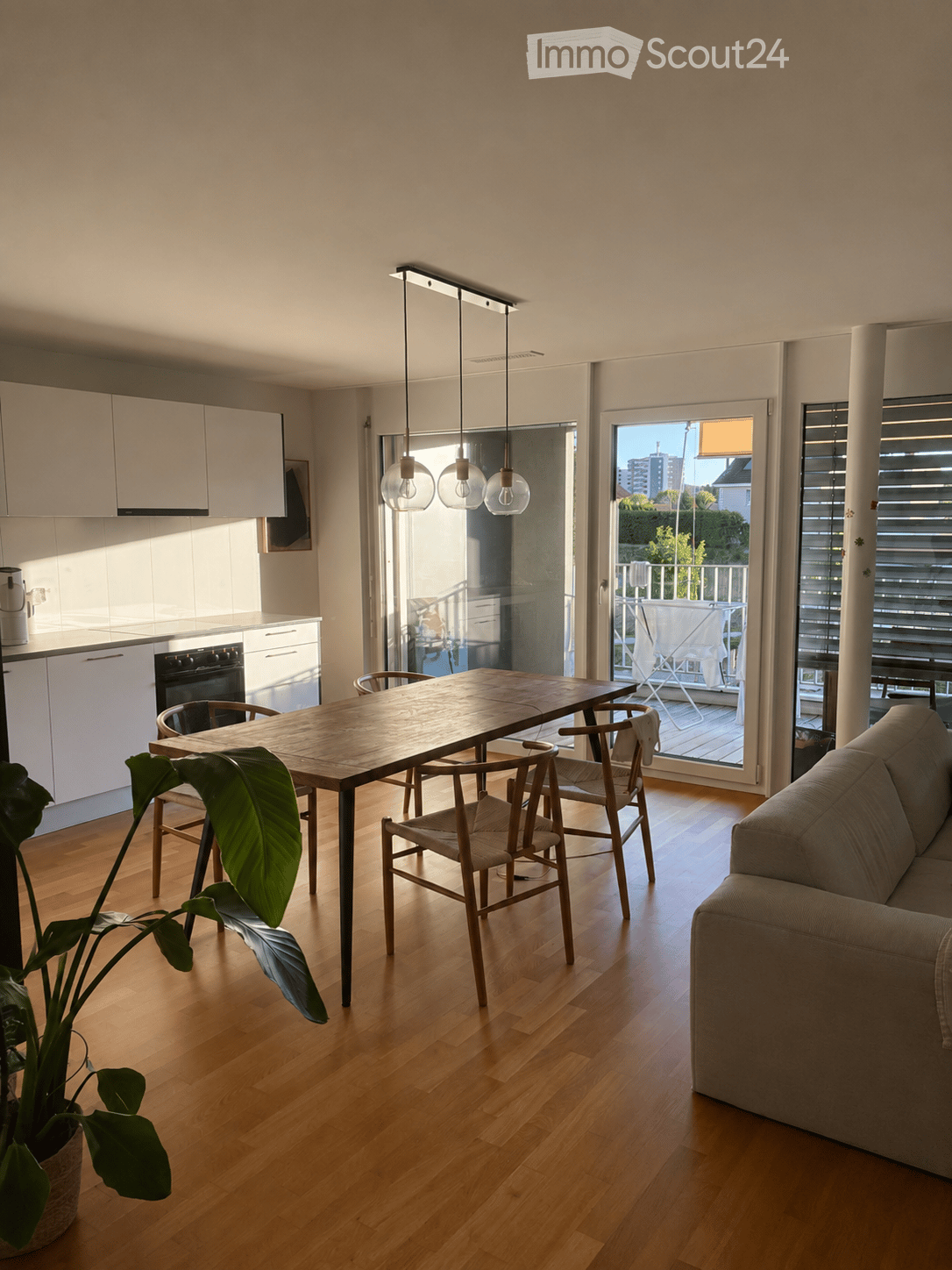 Bright, open-plan living and dining area with wooden flooring, a sofa, a dining table with chairs, a kitchen area with cabinets and an oven, pendant lights, and a potted plant. Large windows and sliding doors lead to a balcony.
