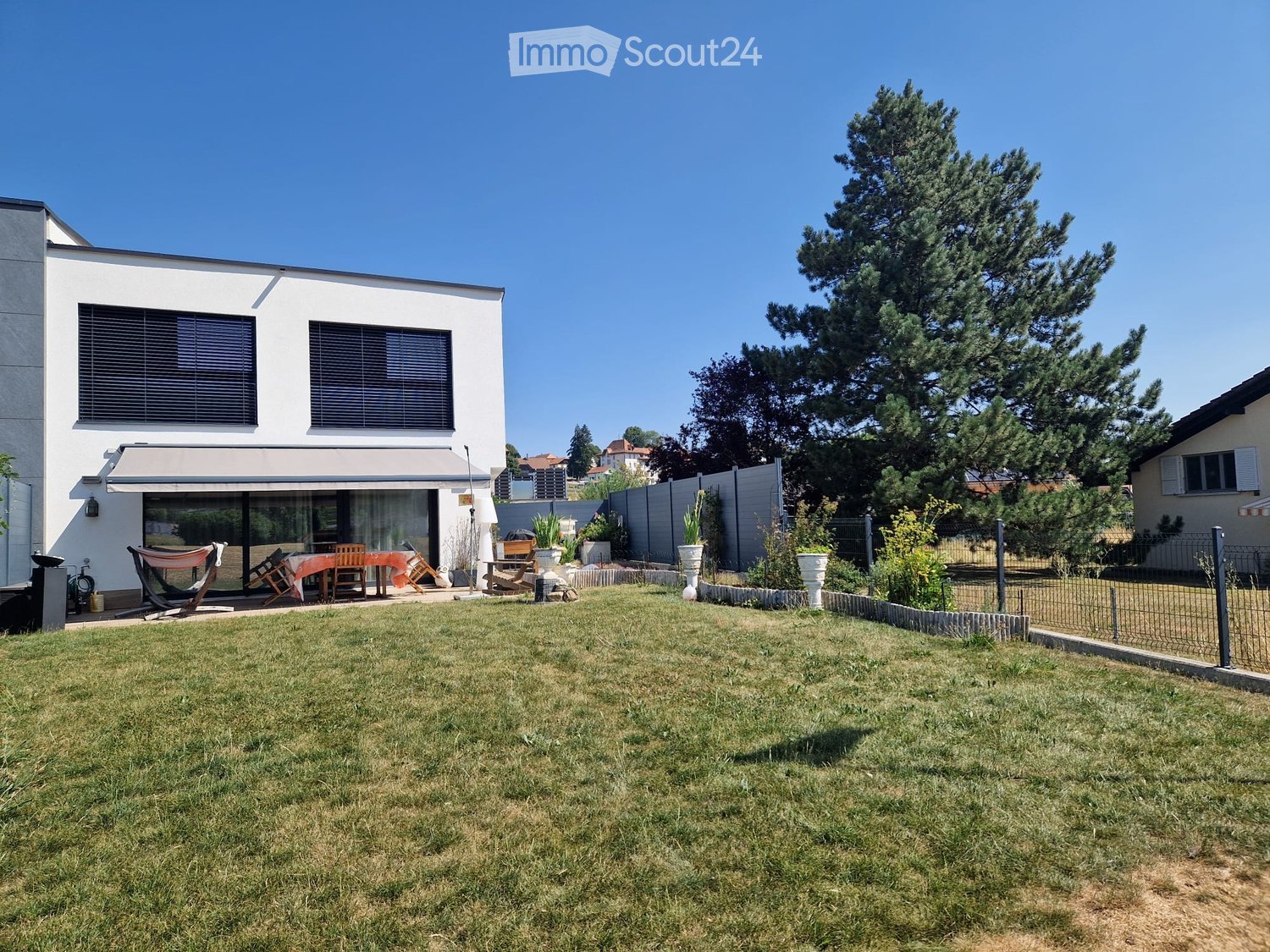 Lawn with a modern house in the background, featuring a patio with outdoor furniture, including chairs and a table, surrounded by trees and a fence.