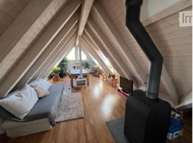 Cozy attic space with wooden beams, exposed wooden floor, grey sofa, coffee table, dining area with table and chairs, television, and a fireplace.