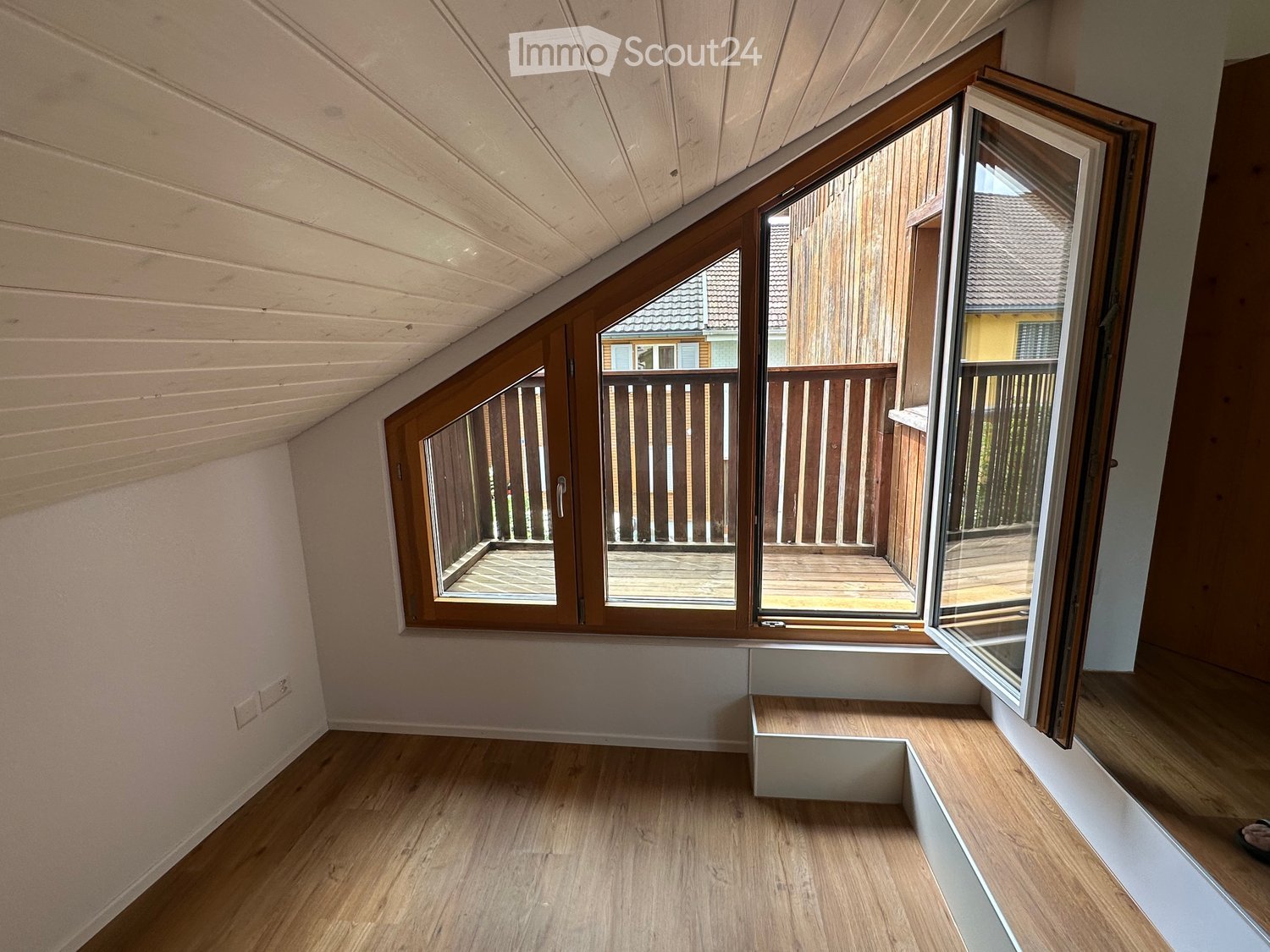An empty attic room with wooden floors, white walls, and large windows opening to a wooden balcony. The ceiling has exposed wooden beams.
