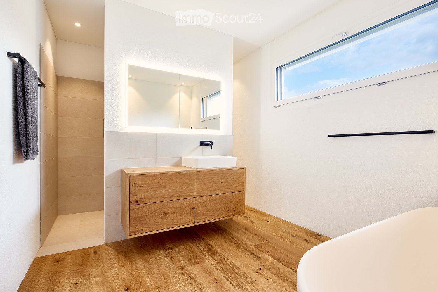 Modern bathroom with wooden vanity, white sink, large mirror, and a towel hanging on the wall. The space features light-colored tiles and a large window letting in natural light.