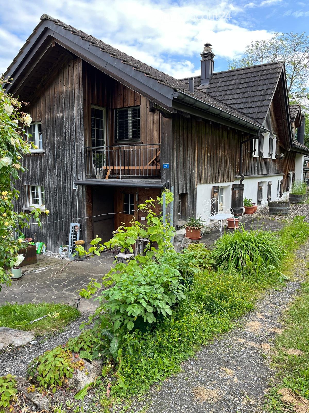 Traditional wooden house with a steep roof, balcony, and surrounding garden with potted plants and outdoor furniture.