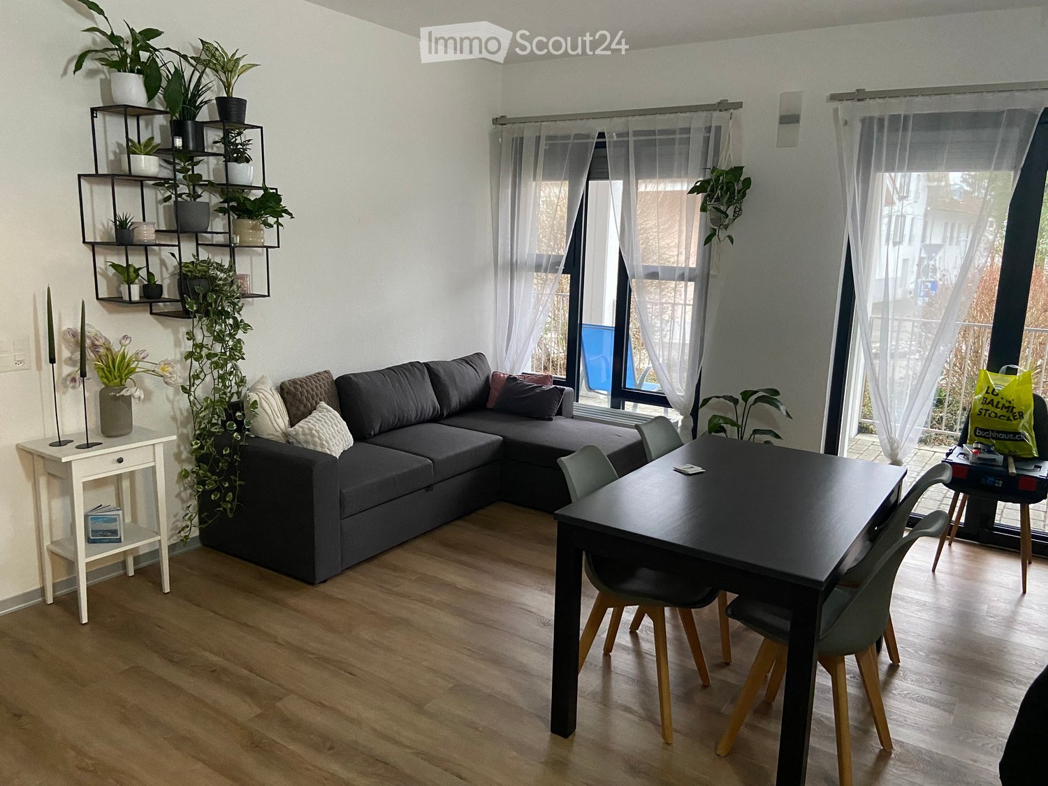Modern living room with a grey sectional sofa, a black dining table with four chairs, a white side table with a vase and candles, and a black shelf with potted plants. Large windows with sheer curtains let in natural light.