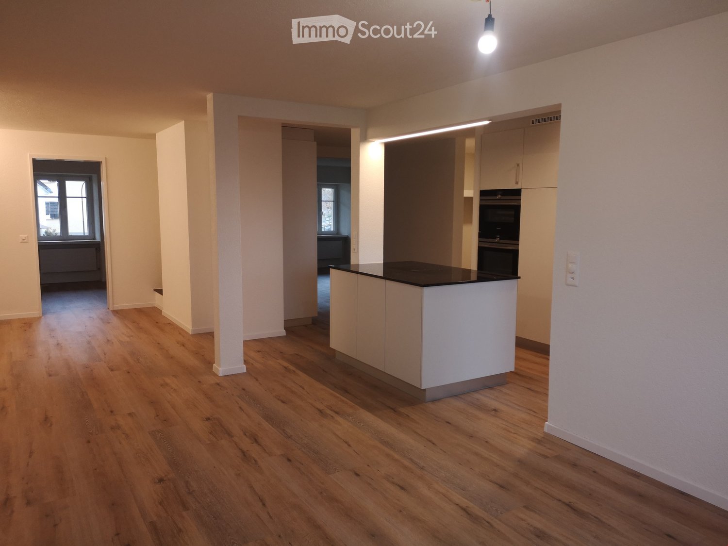 Empty kitchen with light wooden flooring, white walls, and a white kitchen island with a black countertop. The space is well-lit with natural light from windows and a hanging light fixture.