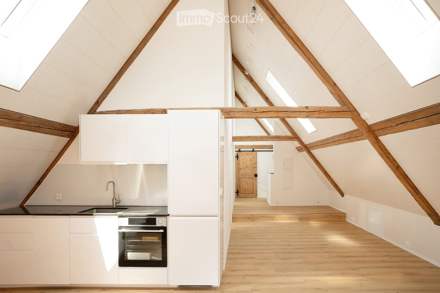 Modern kitchen with white cabinets, stainless steel sink, and built-in oven, featuring exposed wooden beams and skylights.