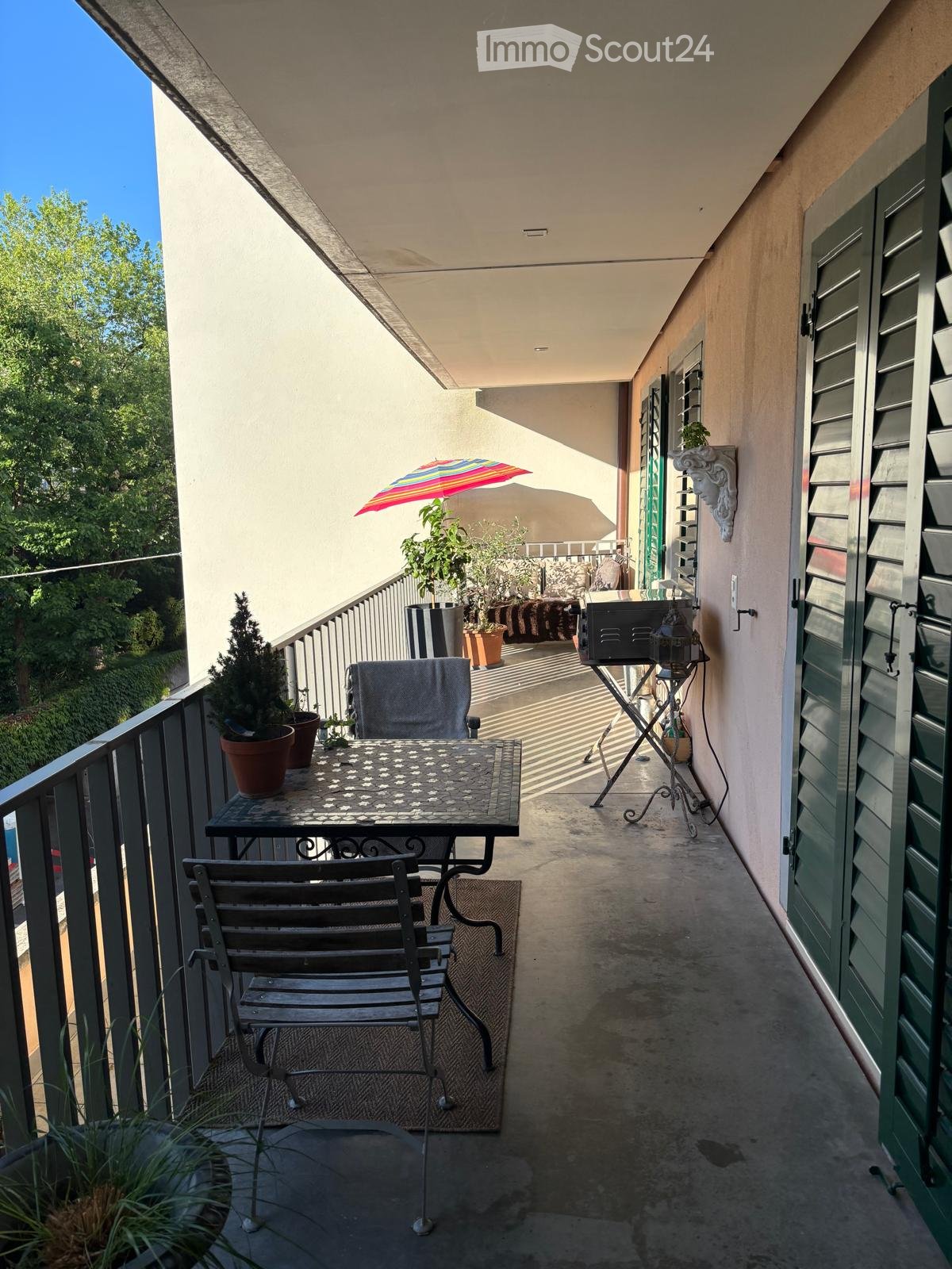 Balcony with a small table, chairs, potted plants, and an umbrella providing shade.