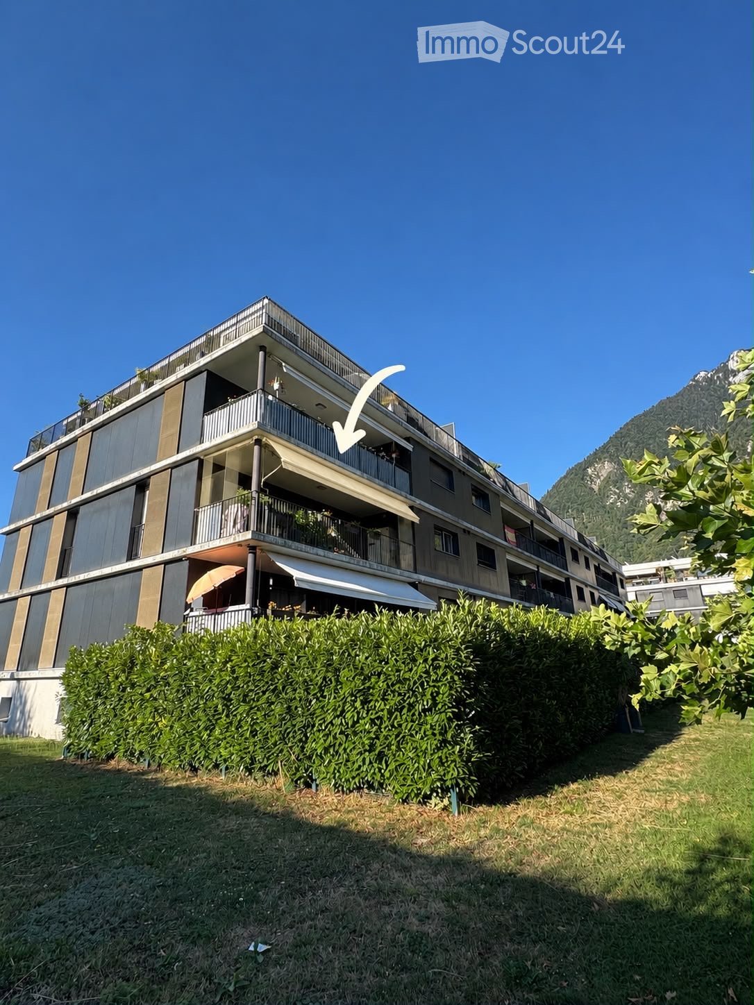 Modern multi-story apartment building with balconies, surrounded by greenery and a mountain view.