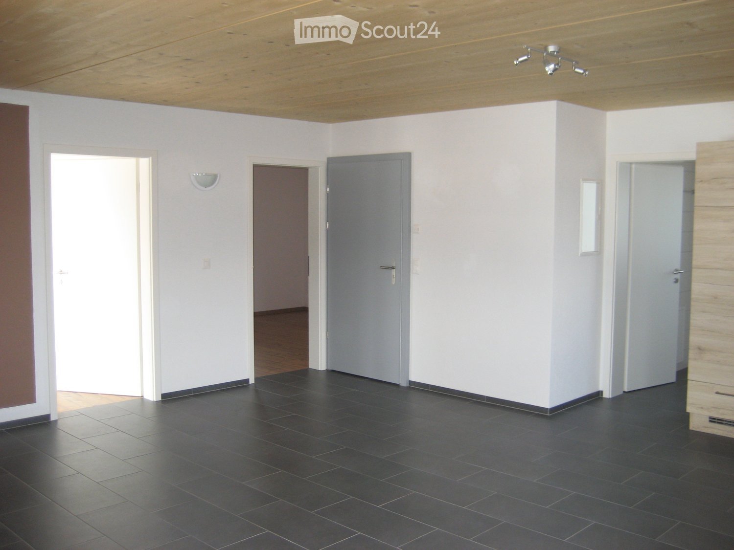 Empty room with white walls, dark tiled floor, wooden ceiling, and several doors leading to other spaces.