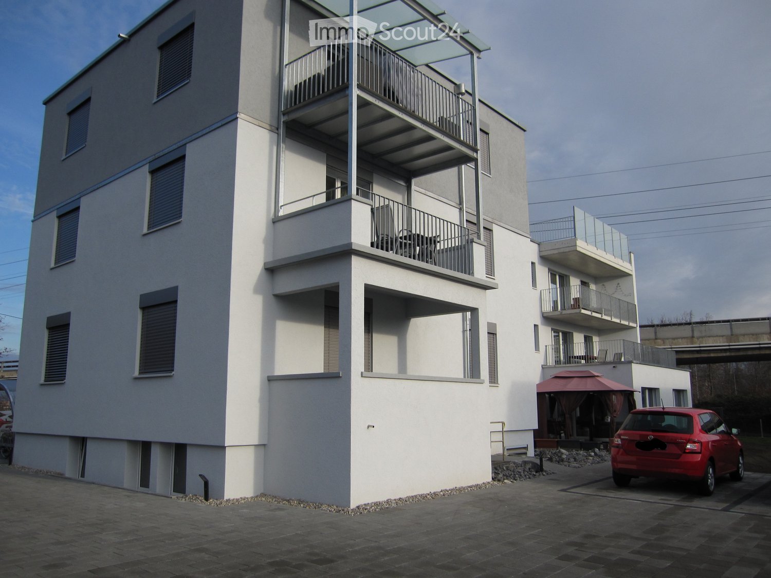 Modern multi-story building with white facade, multiple balconies, and a red car parked nearby.