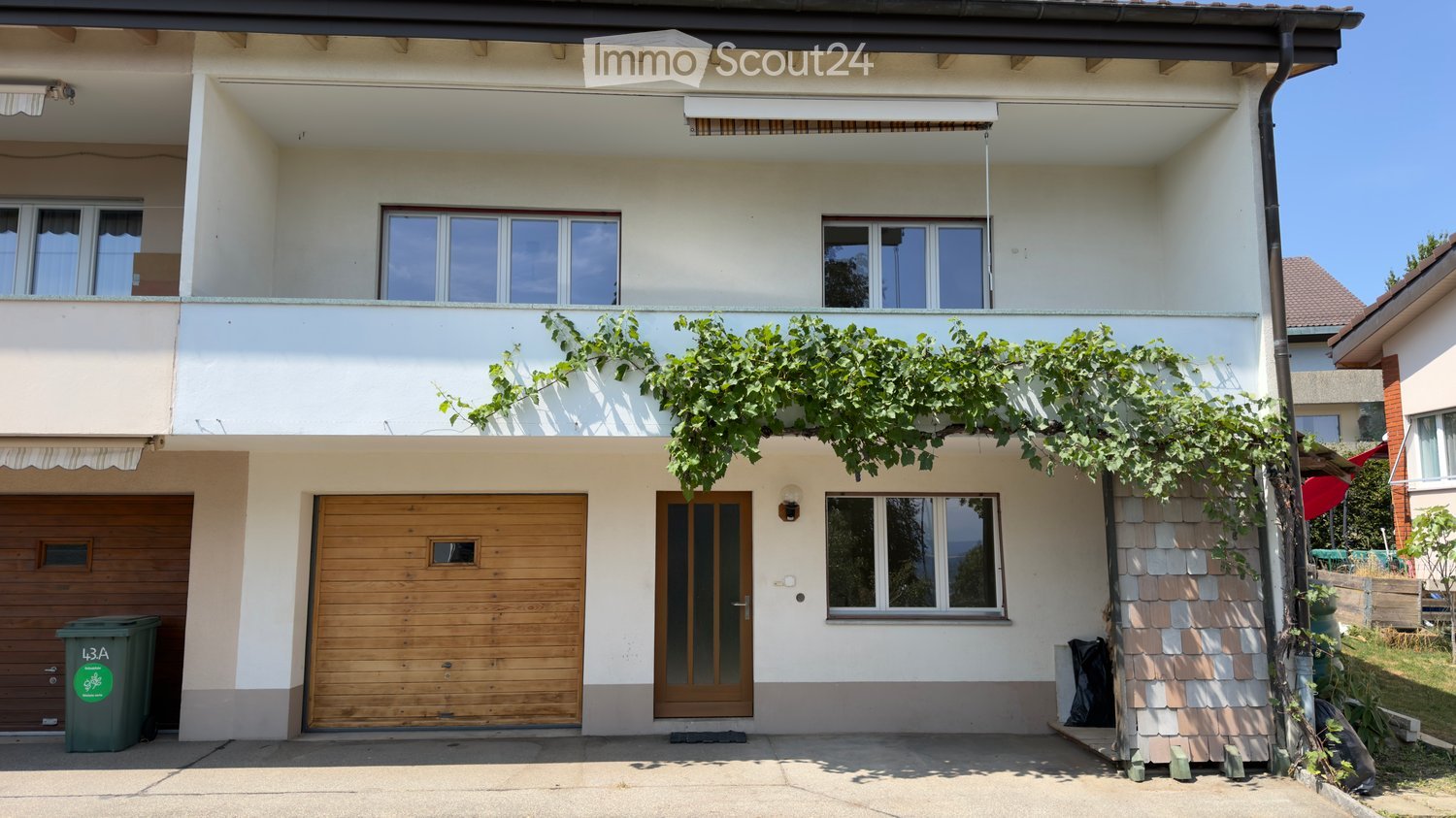 Two-story house with white exterior, wooden garage door, and balcony with green vines. Features multiple windows and a covered entrance area.