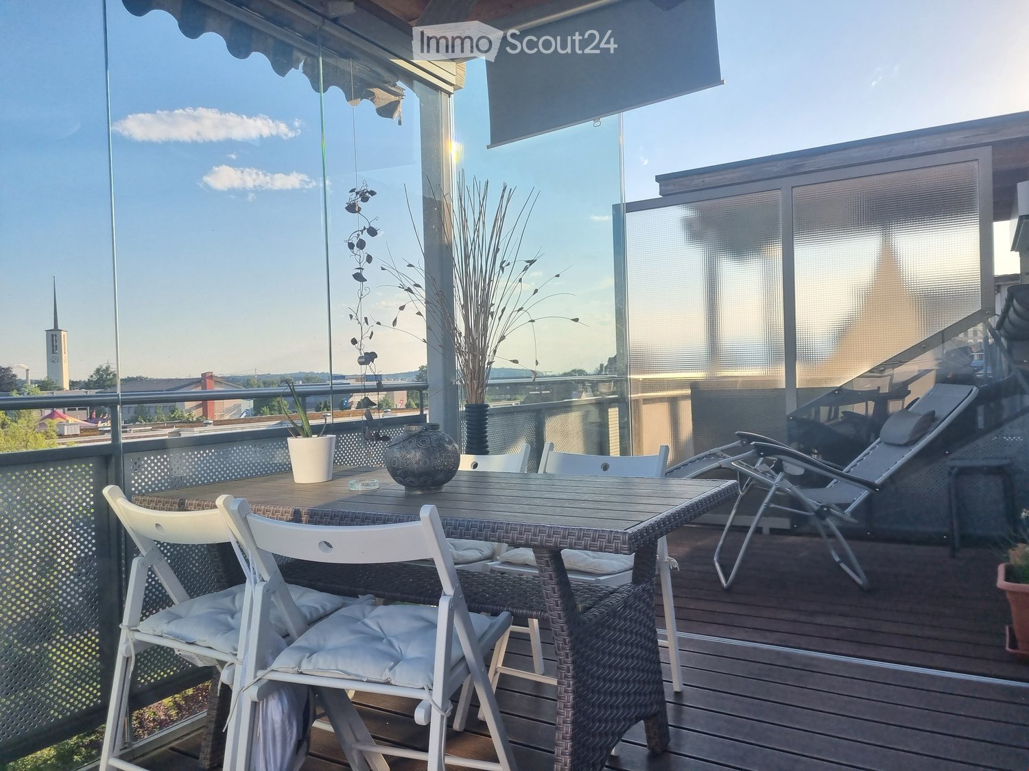 Balcony with wooden decking, glass railing, outdoor table with chairs, lounge chair, decorative vases, and potted plants.