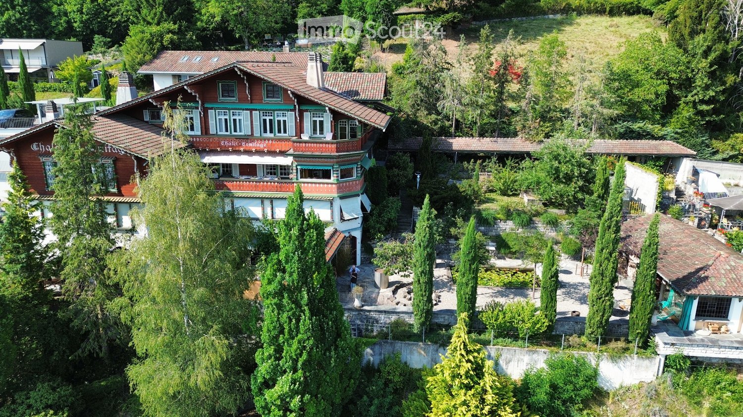 Aerial view of a large, multi-story house with red and white exterior, surrounded by lush greenery and trees. The house features multiple windows, a prominent roofline, and a well-maintained garden area with pathways and outdoor seating.