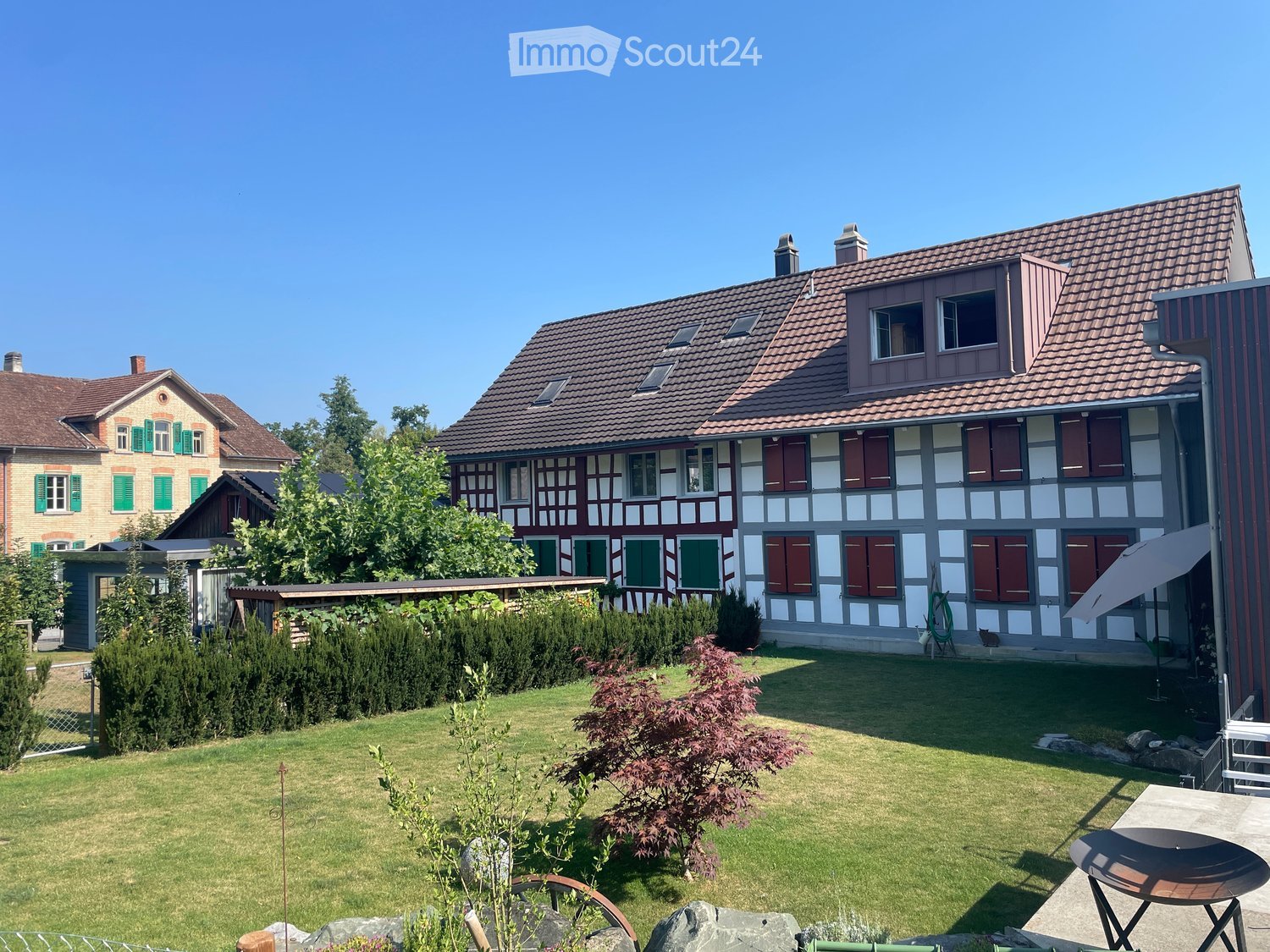 Well-maintained garden with a lawn, shrubs, and a small patio area featuring a table, chairs, and an umbrella. The house in the background has a traditional half-timbered design with red shutters and a tiled roof.