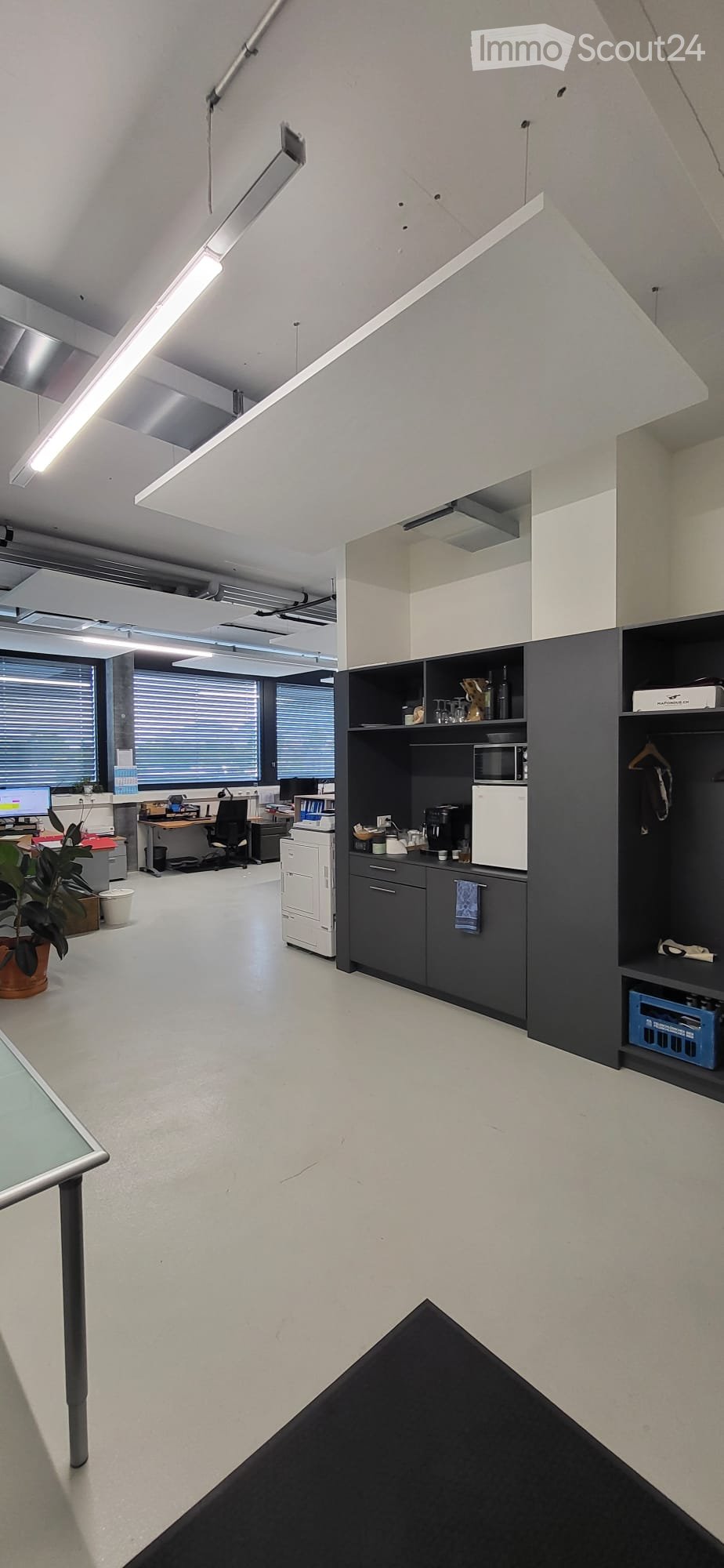 Modern office space with white walls, large windows, and a small kitchenette area. Includes desks, chairs, a printer, a microwave, and a coffee maker.