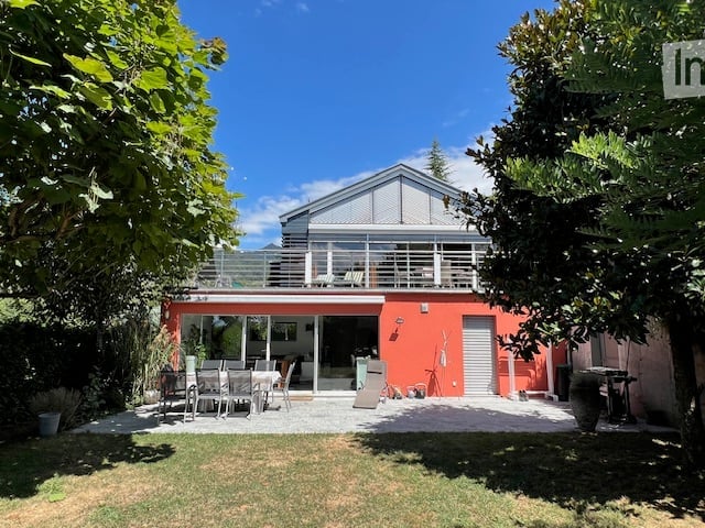 Modern orange house with a large balcony and outdoor dining area, surrounded by greenery and trees.