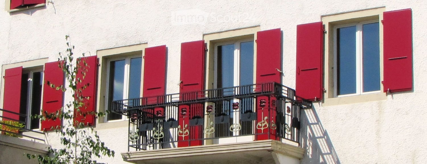 Balcony with black metal railing and decorative elements, red shutters, and large windows on a white building facade.