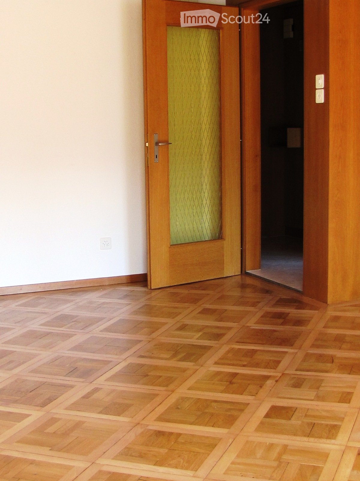 A narrow hallway with wooden panel walls and a frosted glass window with a wooden frame. The walls are light-colored with a geometric pattern, and the floor is not visible.