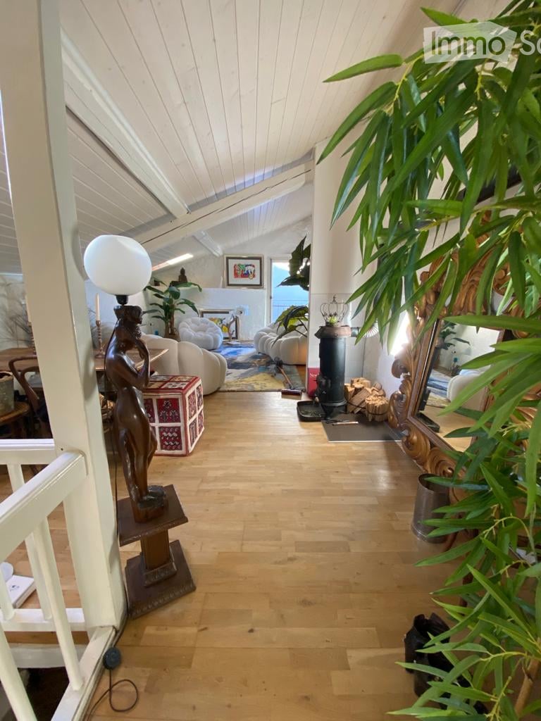 Bright, open living space with wooden floors, white paneled ceiling, fireplace, sofa, chairs, table, lamp, and decorative plants.