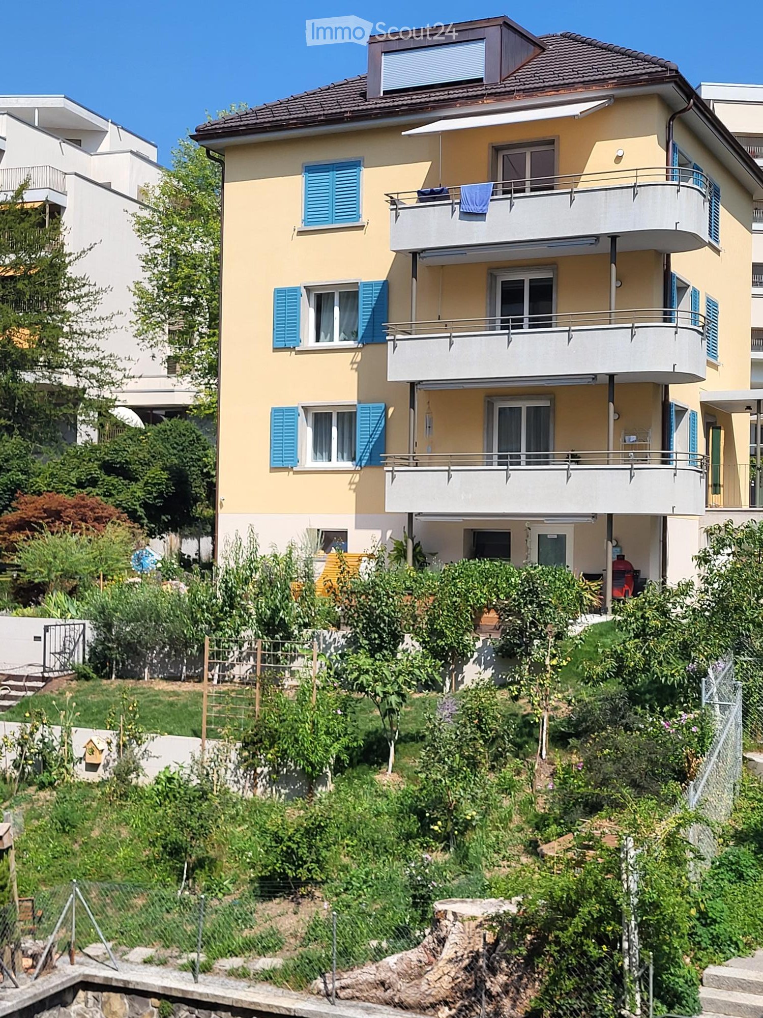 Two-story house with yellow walls, blue shutters, multiple balconies, and a well-maintained garden on a raised ground floor.