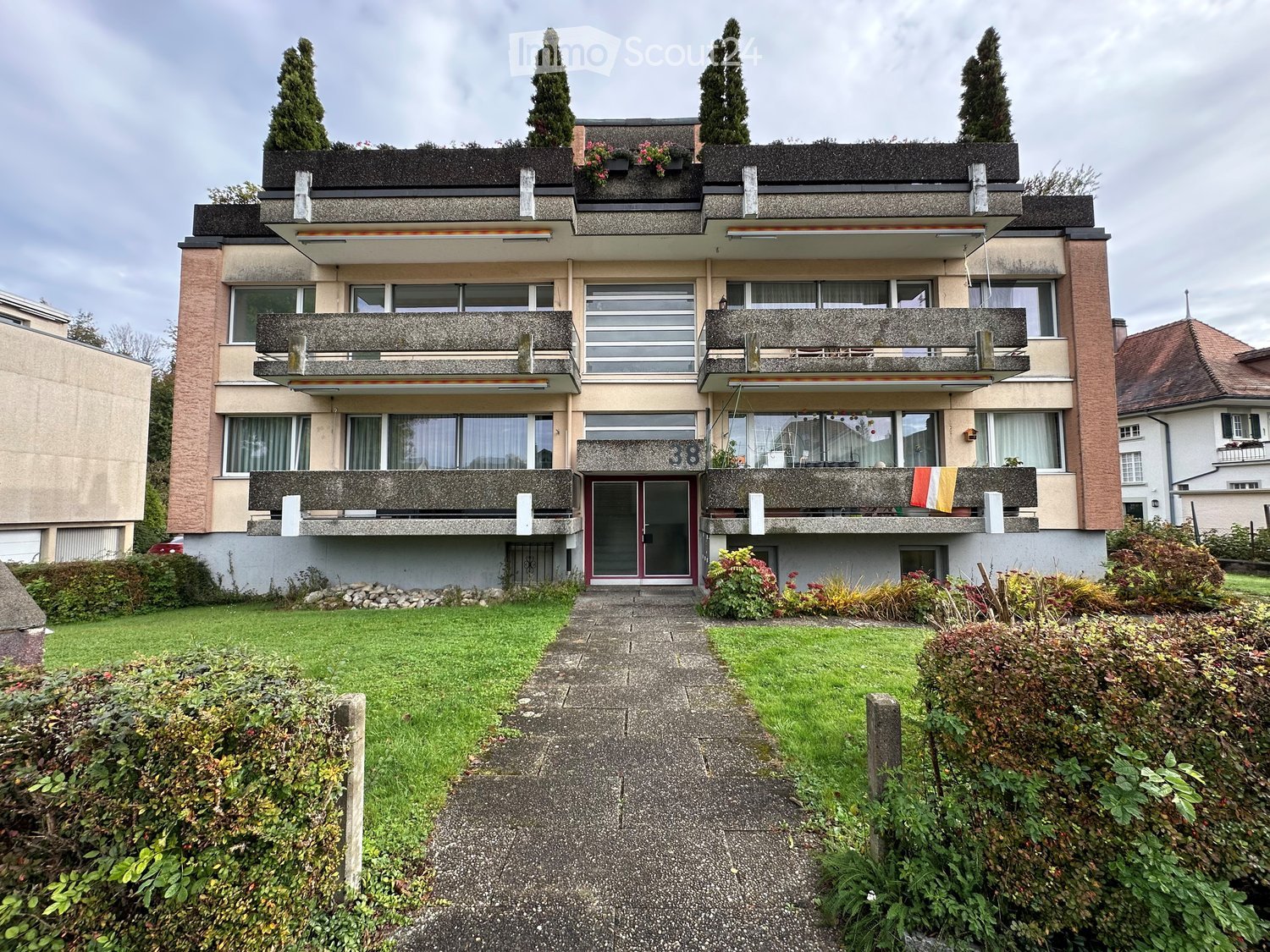 A three-story residential building with balconies, large windows, and a well-maintained garden in front.