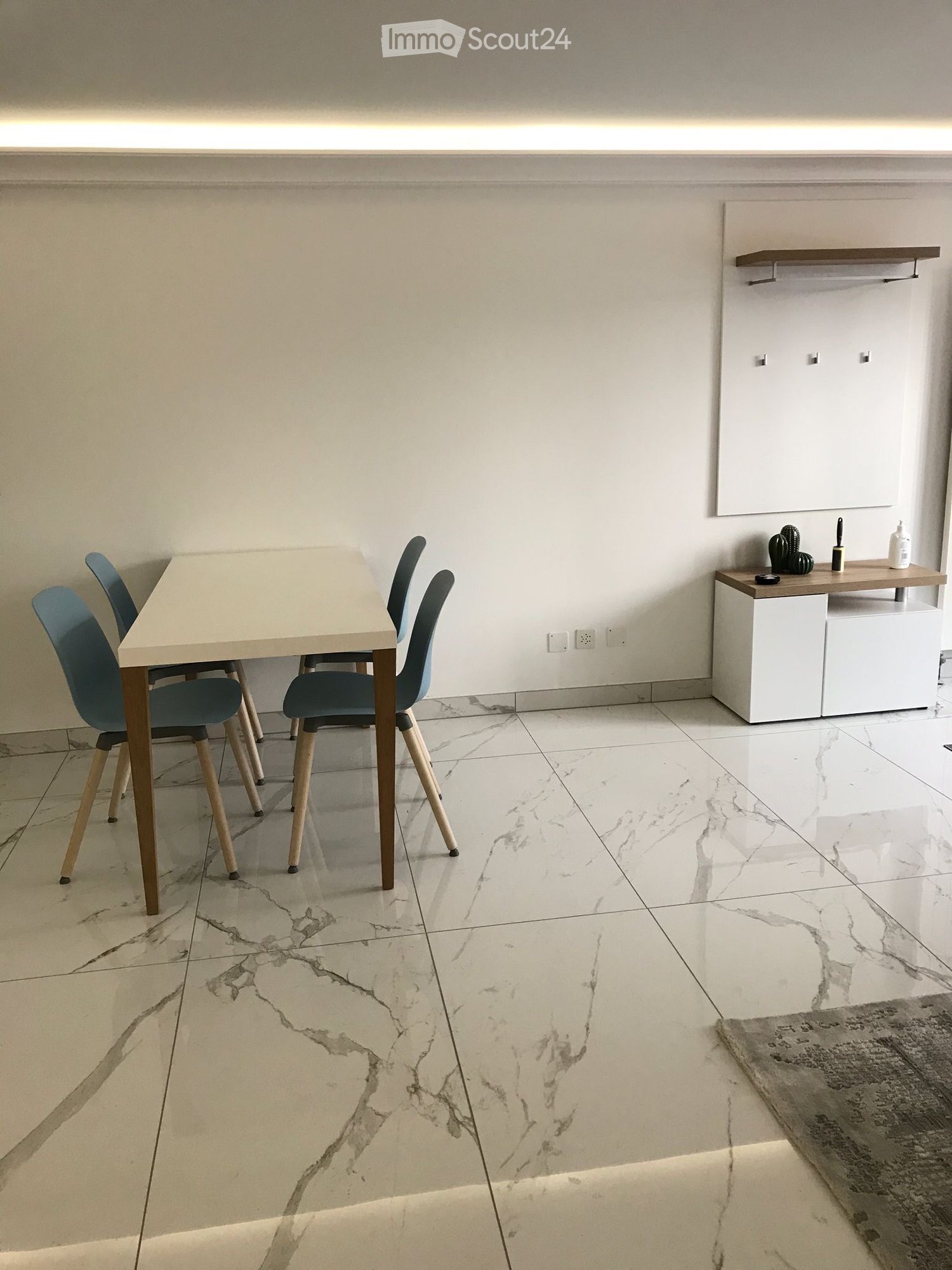 Dining area with a light-colored table, four modern chairs, a white cabinet, and a small shelf with decorative items.