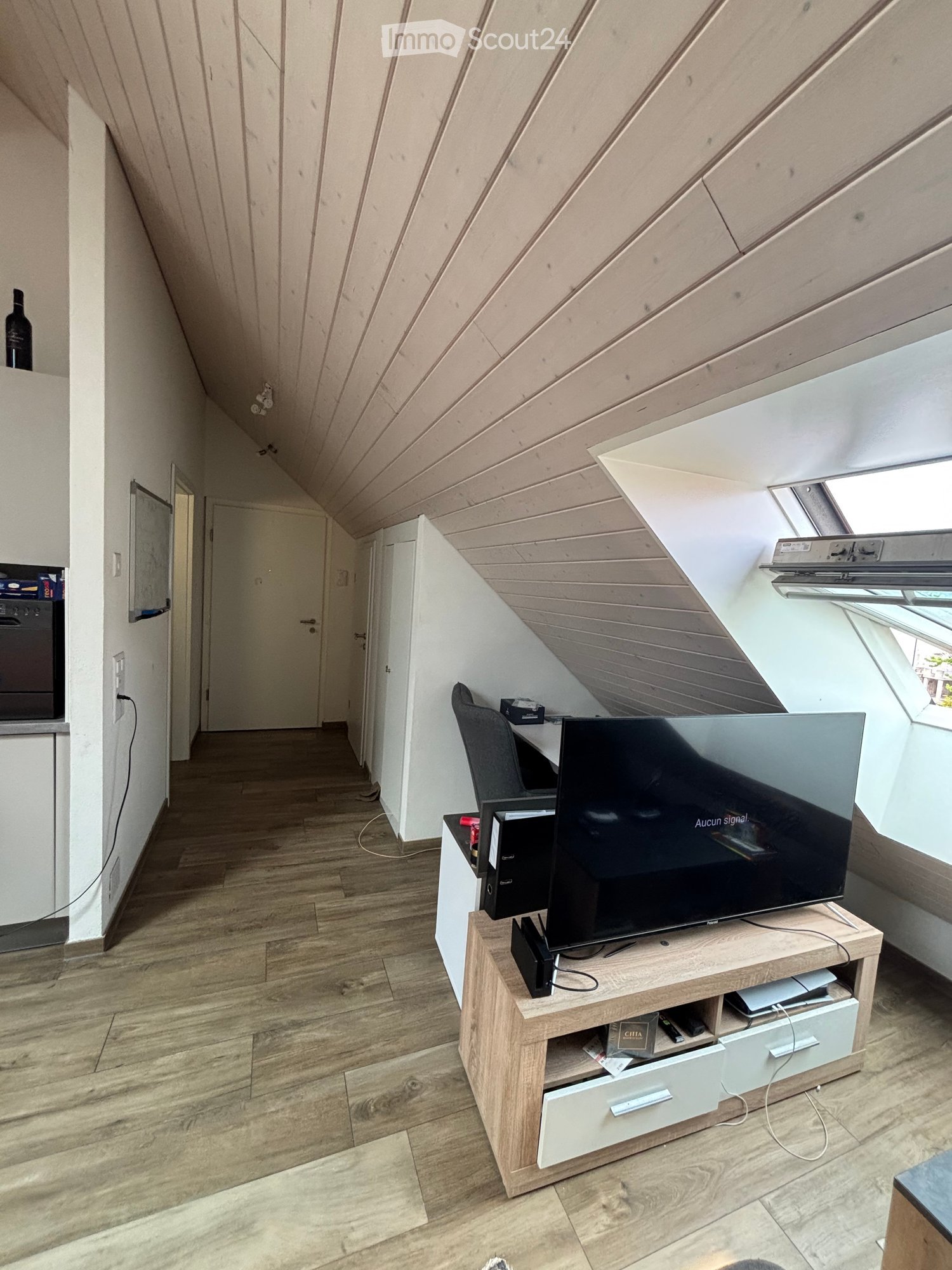 Modern living room with wooden flooring, slanted wooden ceiling, TV stand with flat-screen TV, chair, desk, and a bottle on a shelf.