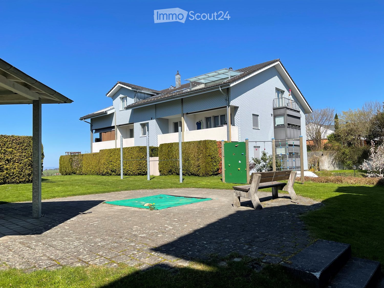 A modern two-story house with white siding and multiple windows, surrounded by a well-maintained garden with a climbing wall, a wooden bench, and a small play area.