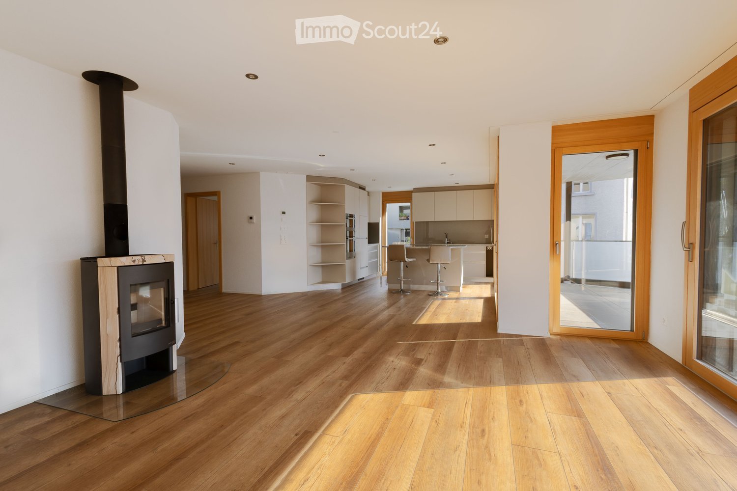 Open-plan living and dining area with wooden flooring, a modern fireplace, and a small kitchen area with cabinets and stools.