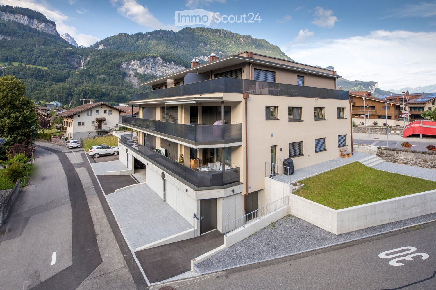 Modern multi-story building with balconies, surrounded by a parking area and scenic mountain backdrop.