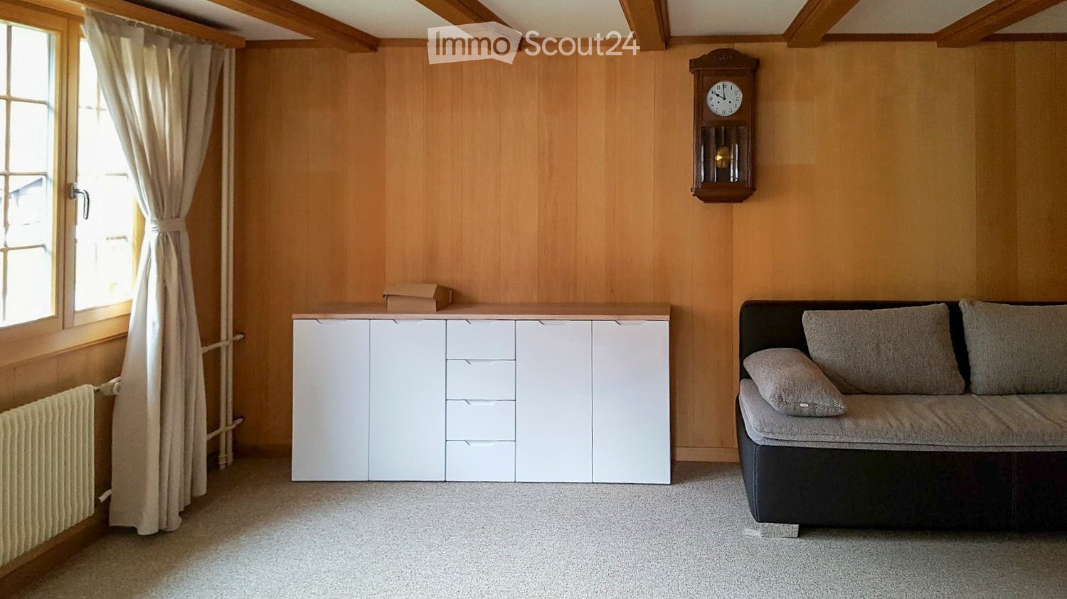 A cozy living room with a dark-colored sofa, a white cabinet, and a wall-mounted wooden clock. The room features wooden panel walls, a large window with curtains, and a radiator below the window.