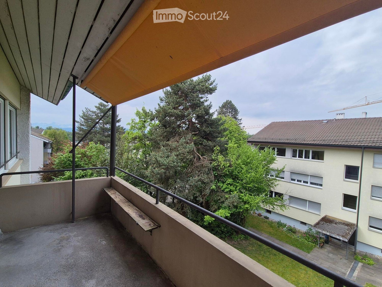 Balcony with a metal railing, overlooking a residential area with trees and neighboring buildings.