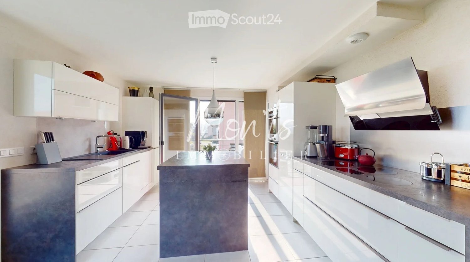 Modern kitchen with white cabinets, grey countertop, central island, and various appliances including a sink, oven, microwave, blender, coffee maker, toaster, and teapot. A pendant light hangs above the island.
