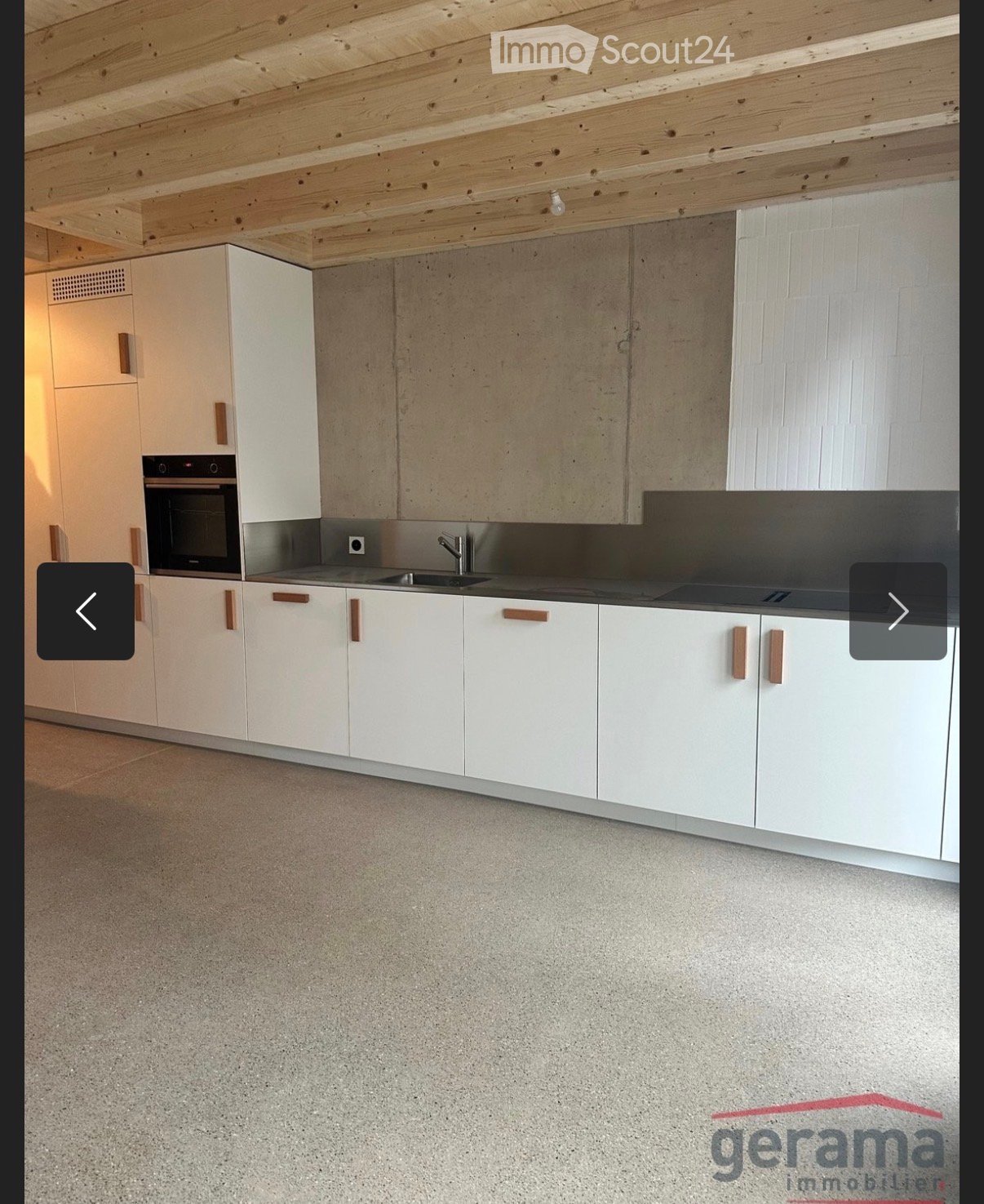 Modern kitchen with white cabinets, a sink, a faucet, and an oven, featuring a concrete backsplash and wooden ceiling beams.