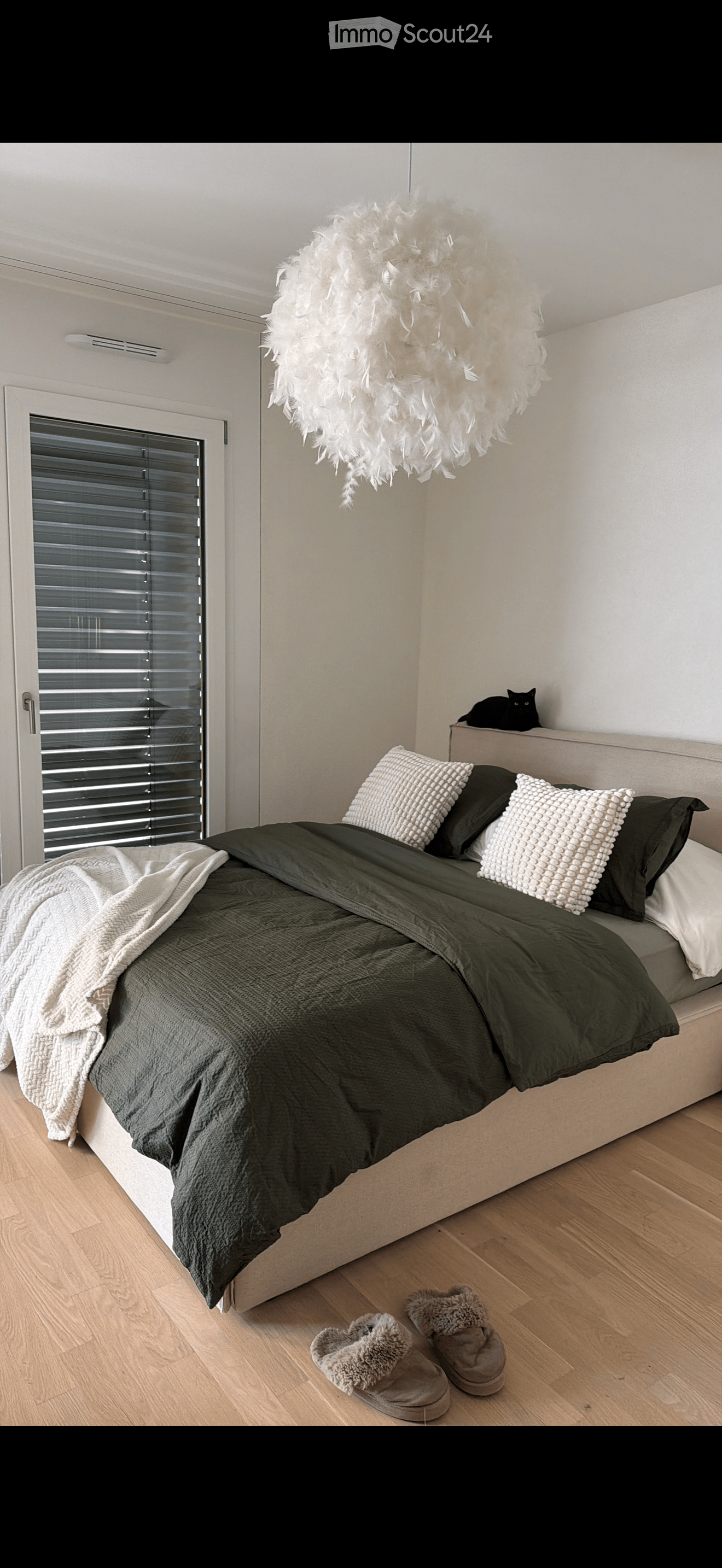 Modern bedroom with a large bed, dark green bedding, white pillows, a fluffy white lamp, a black cat on a shelf, and a pair of slippers on the floor.