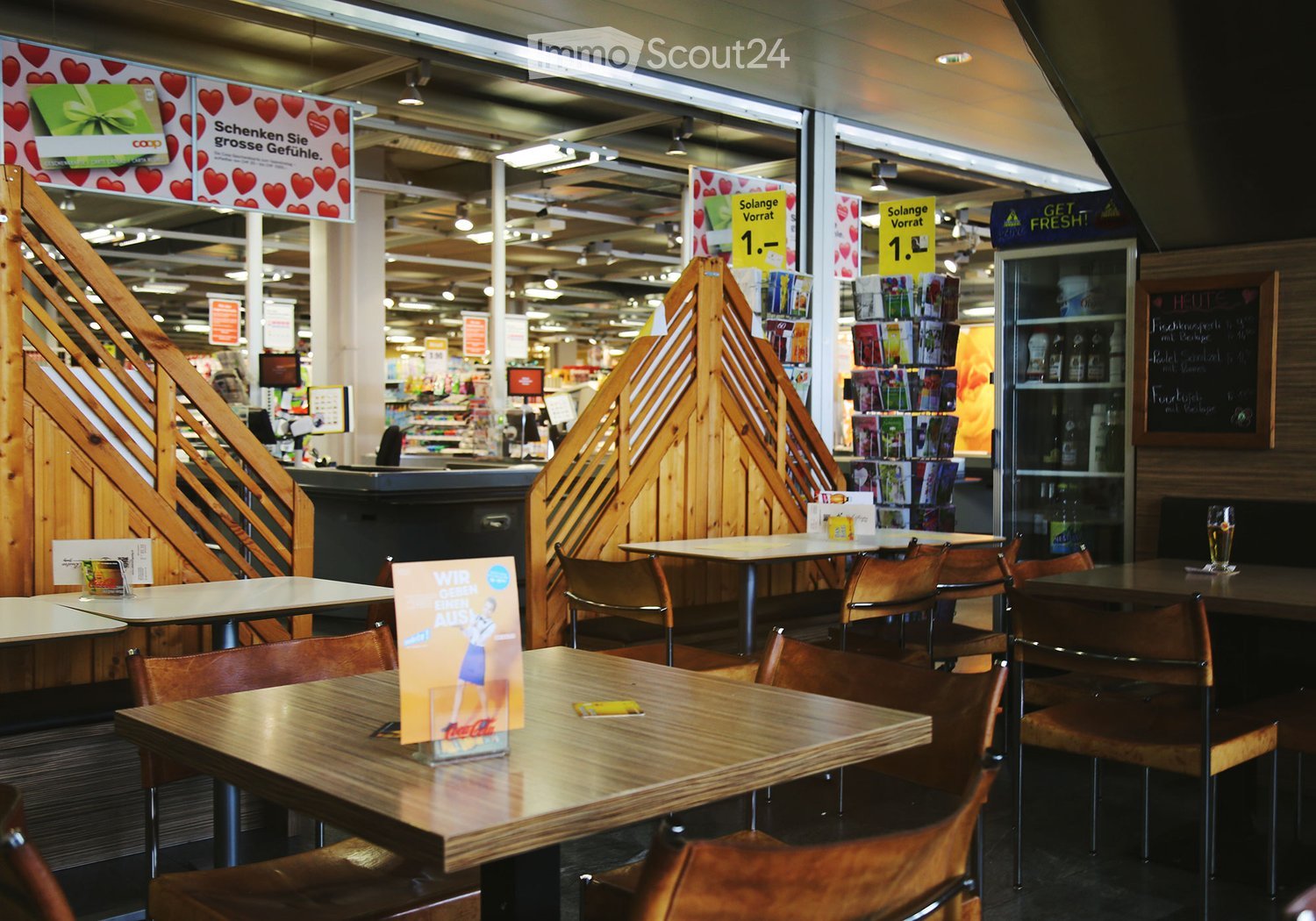 A modern cafeteria area with wooden tables and chairs, a refrigerator, and promotional signs. The space is well-lit with a clean, organized layout.