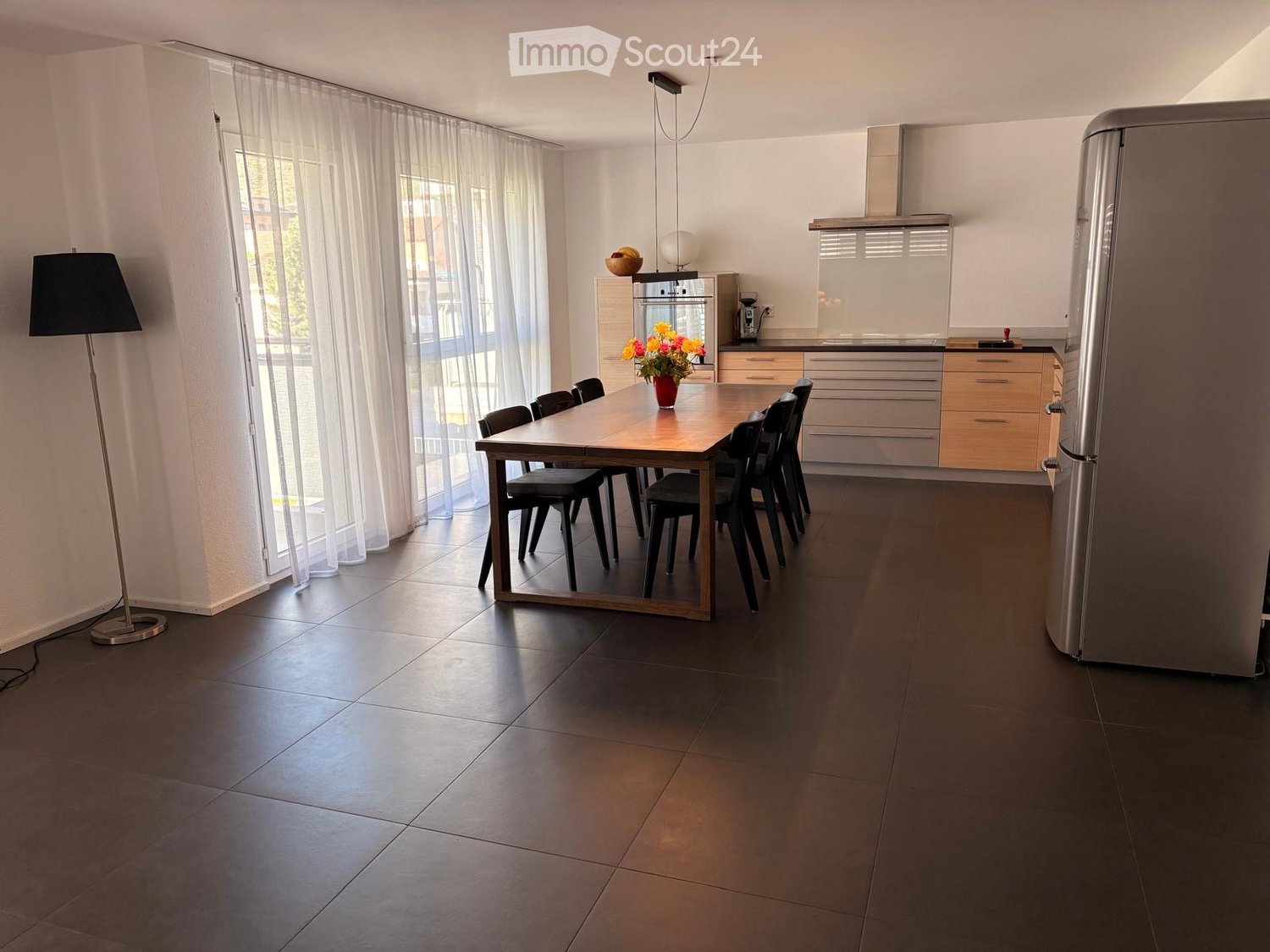 Modern kitchen with a dining area, featuring a wooden dining table, black chairs, a stainless steel refrigerator, and a built-in oven. The space includes a floor lamp and a vase with flowers on the table.