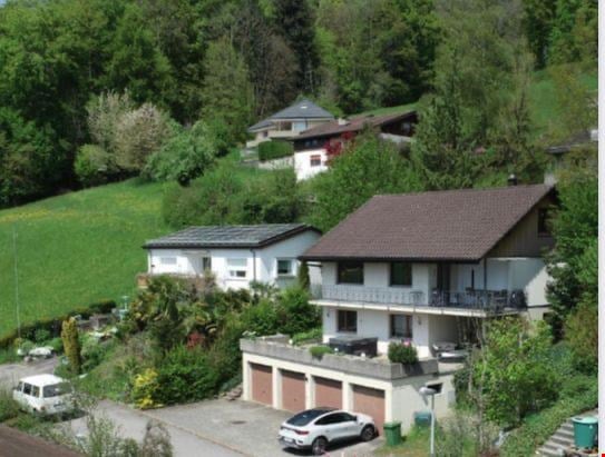 Aerial view of a two-story house with a garage, surrounded by greenery and other houses on a hillside.