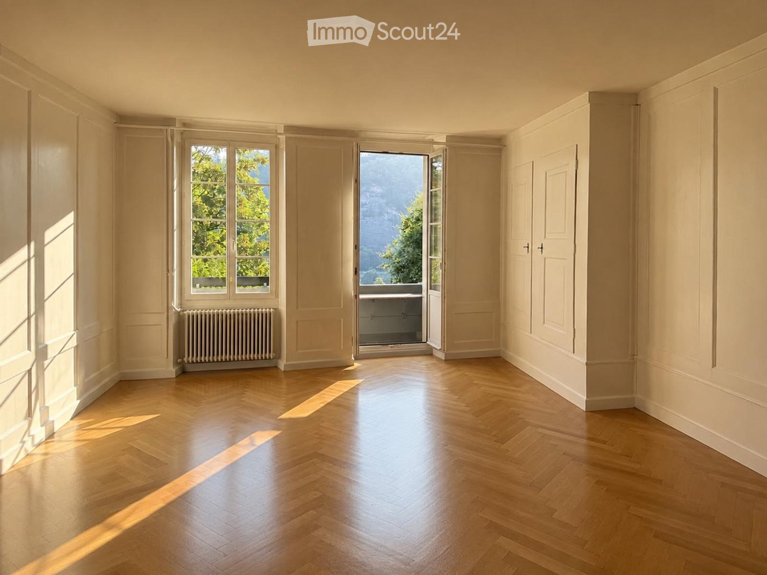 Empty room with light wooden flooring, white walls, large windows, and a radiator. No furniture or equipment present.