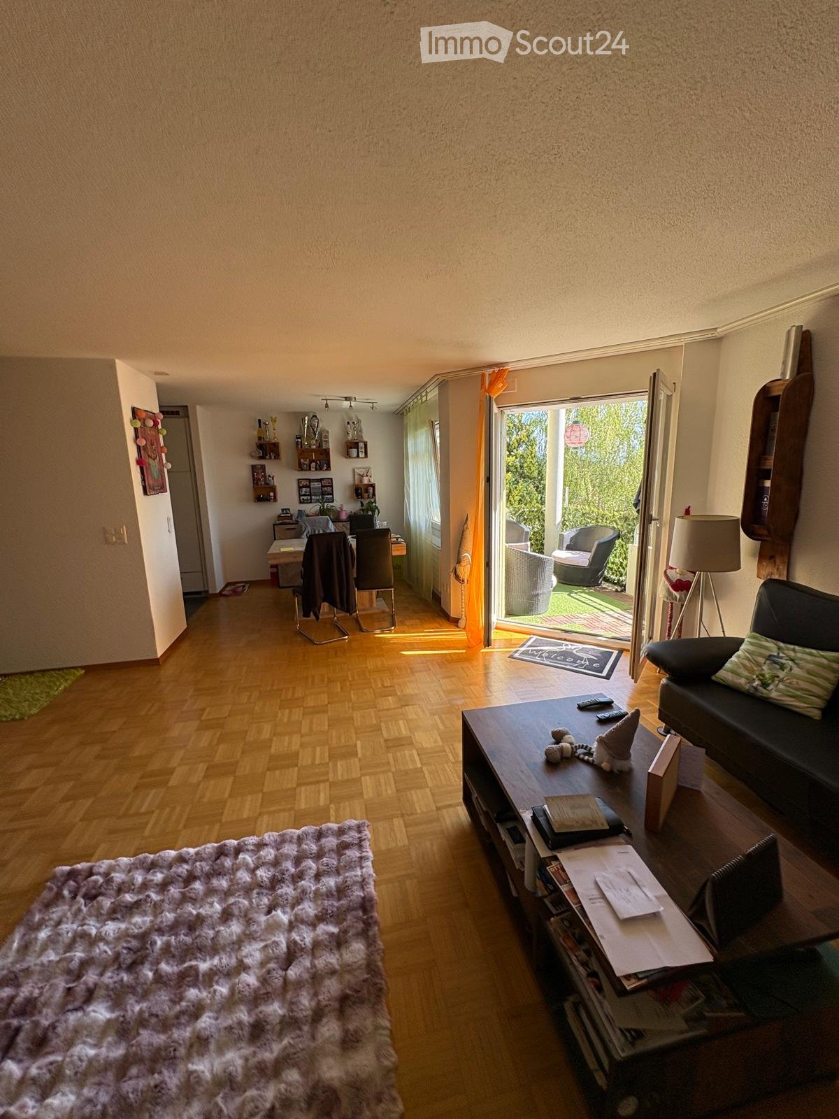 Living room with wooden flooring, a large sofa, coffee table, and a dining area with chairs and a table. There is a balcony with outdoor seating visible through the open sliding doors.