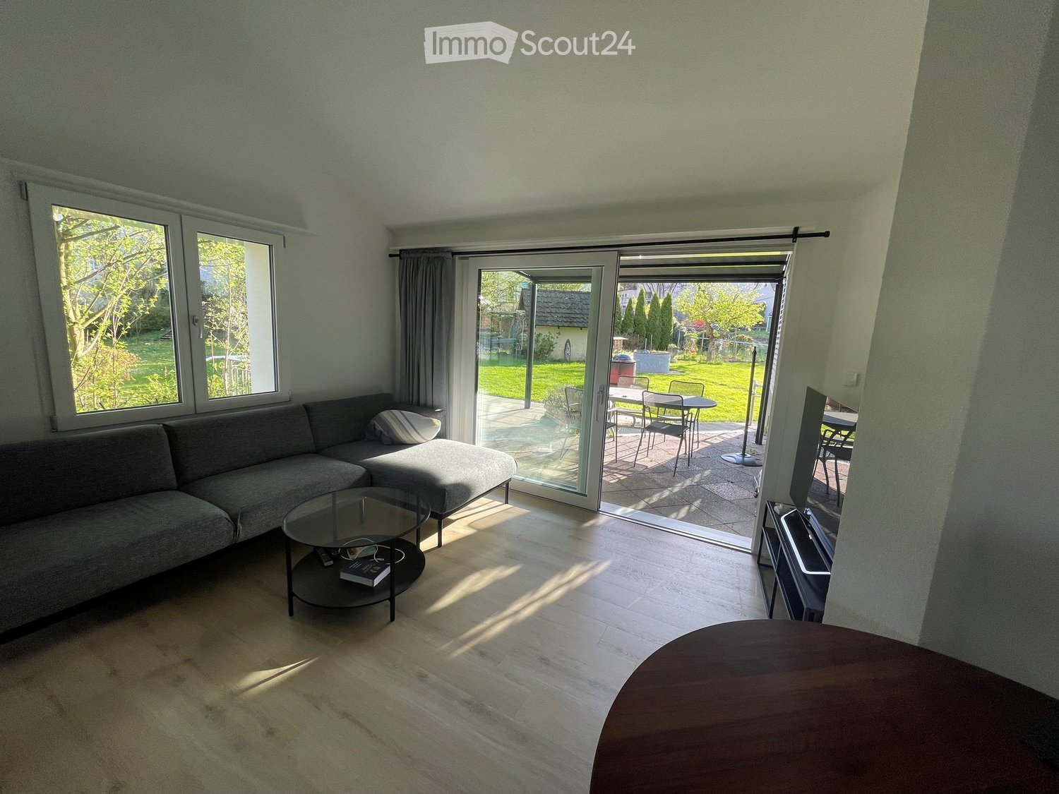 Bright living room with large windows and sliding glass doors leading to a patio. Features a grey sectional sofa, glass coffee table, ottoman, and a TV stand.