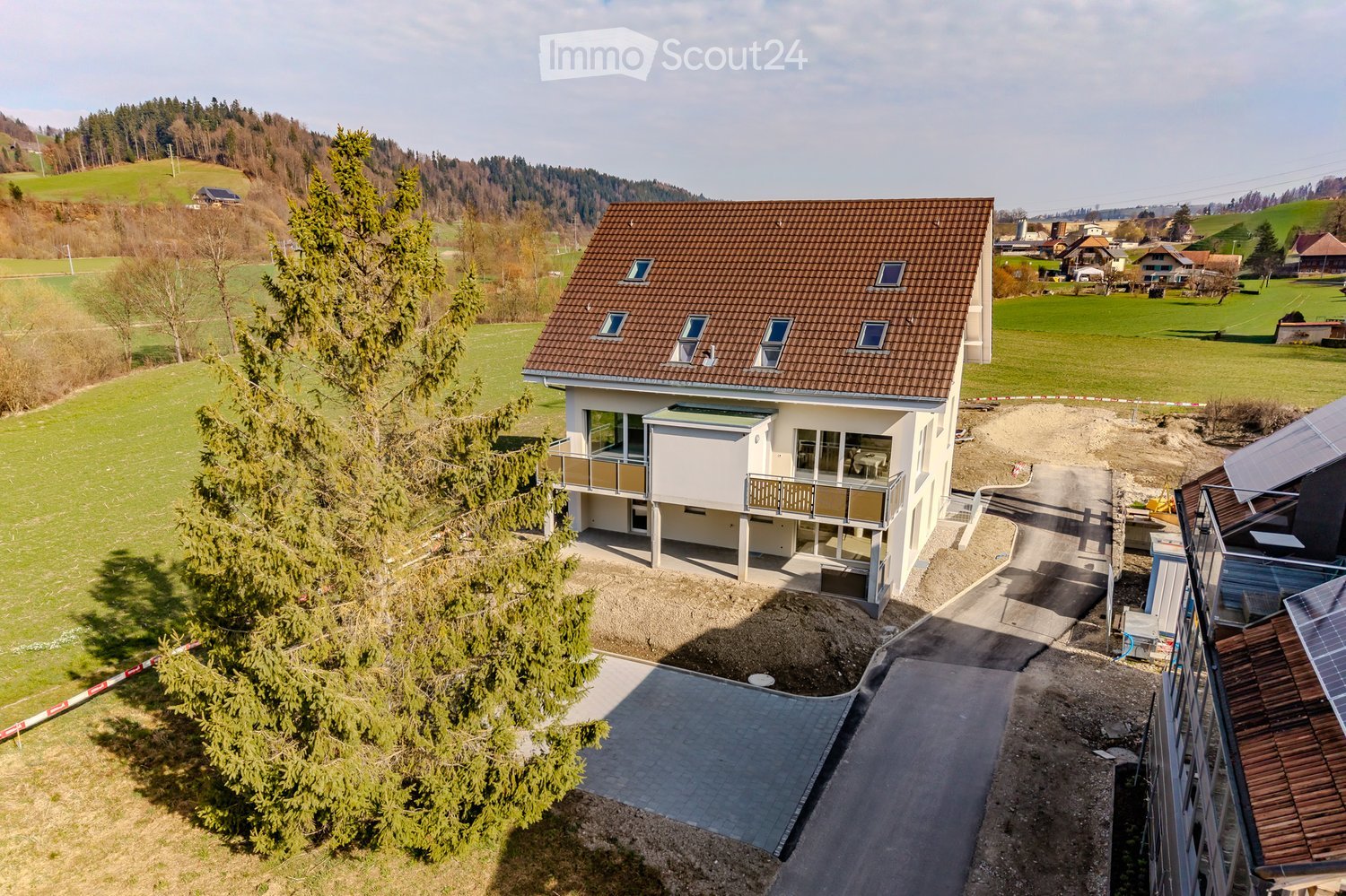 A modern two-story house with a brown tiled roof, large windows, and balconies, situated in a rural landscape with green fields and trees.