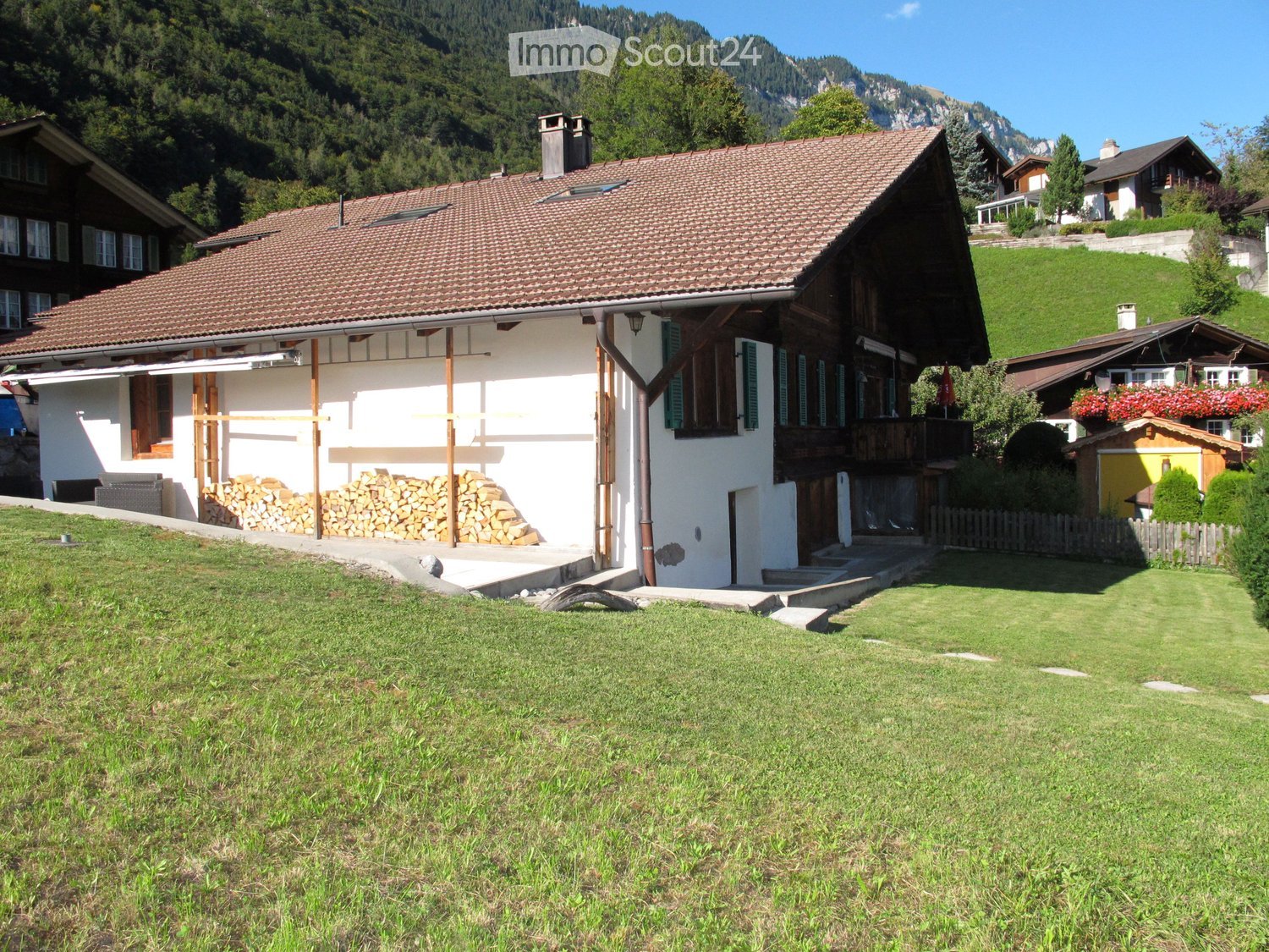 Traditional Swiss house with a tiled roof, wooden balcony, and a stack of firewood near the entrance, surrounded by a grassy lawn and neighboring houses in a mountainous area.