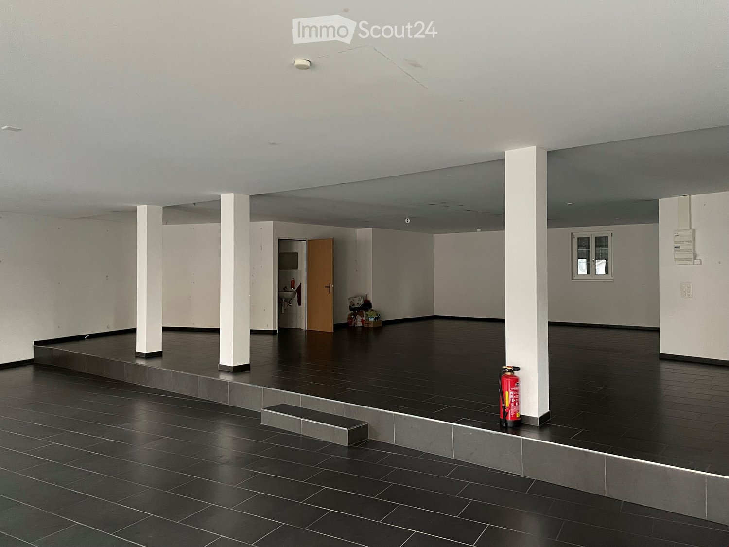 Empty room with dark tiled floor, white walls, and white pillars. A small bathroom area is visible in the background, and a red fire extinguisher is placed near a pillar.