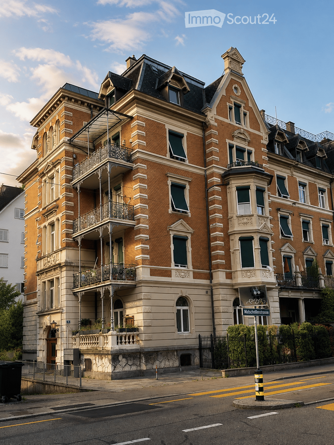 A large, multi-story brick building with ornate architectural details, including balconies, decorative railings, and a prominent roofline. The building features multiple windows with shutters and is situated on a street corner with a visible street sign.