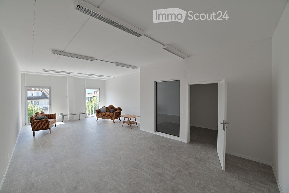 Bright, minimalist room with white walls and concrete flooring, featuring a sofa, small table, and chairs near large windows and a glass door leading to an outdoor area.