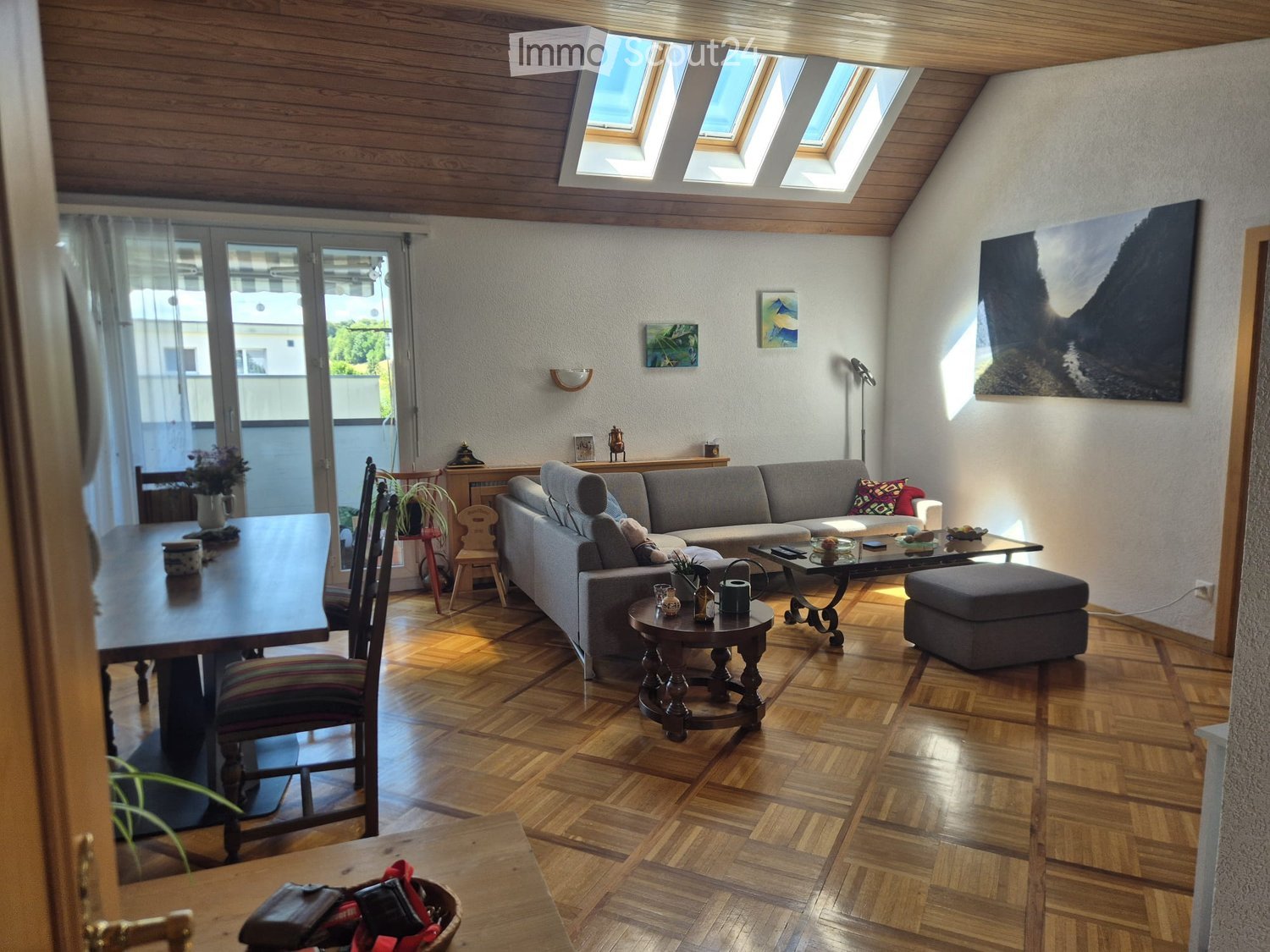 Living room with wooden flooring, grey sofa, coffee table, ottoman, dining table, chairs, and artwork on the walls.